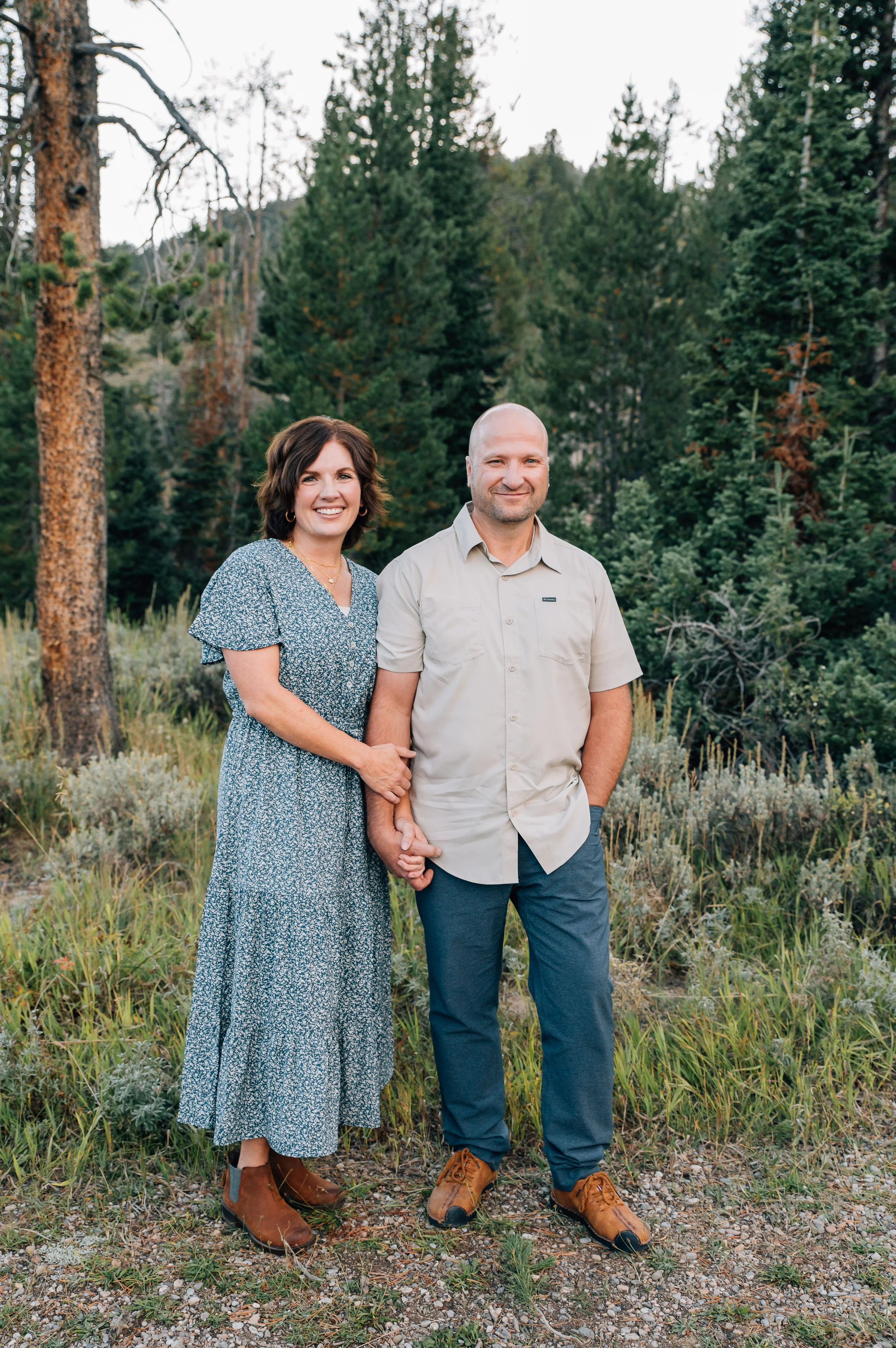 A smiling couple standing outdoors in a forested area holding hands, with trees and greenery in the background.