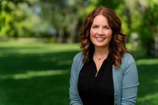 A smiling woman with curly auburn hair standing outdoors in a park with green grass and trees in the background.