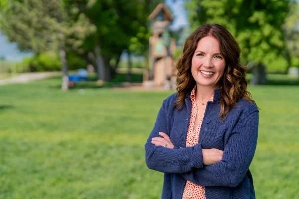 A woman with brown hair smiling and standing with arms crossed in a park with trees and a sculpture in the background.
