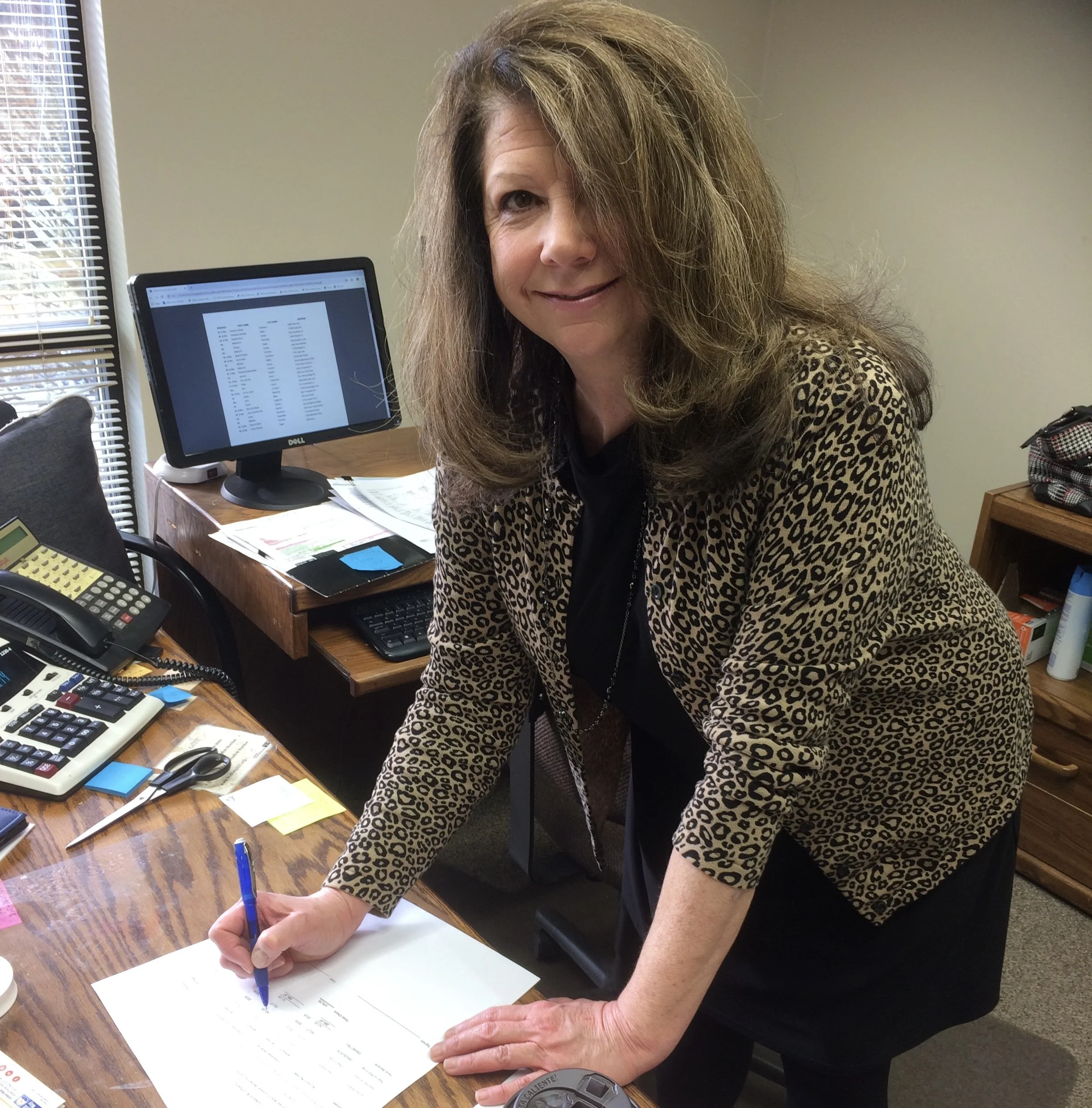 Regina smiling at the camera while writing on a piece of paper at a desk.