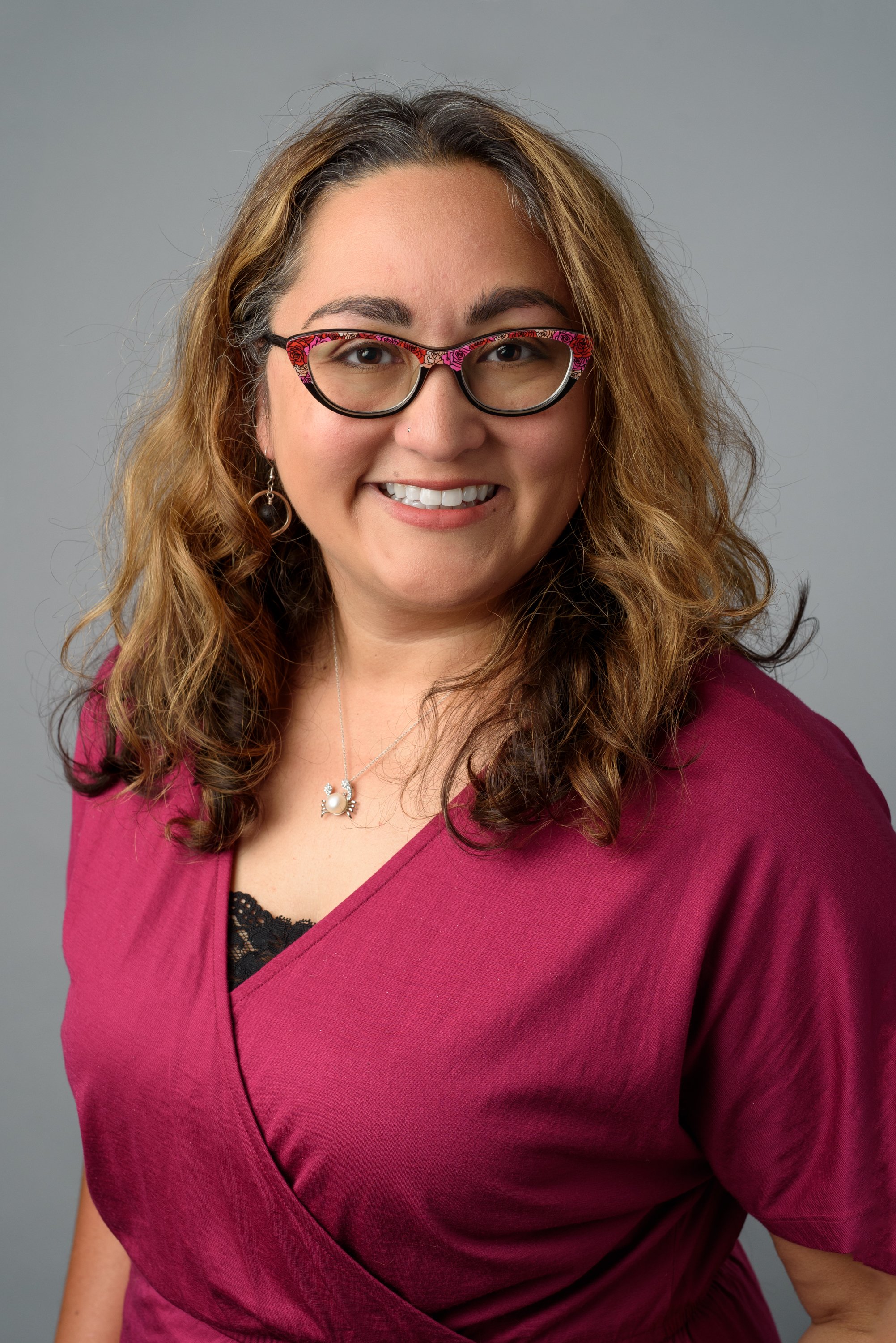 Maya smiling at the camera wearing a red blouse and floral print glasses.