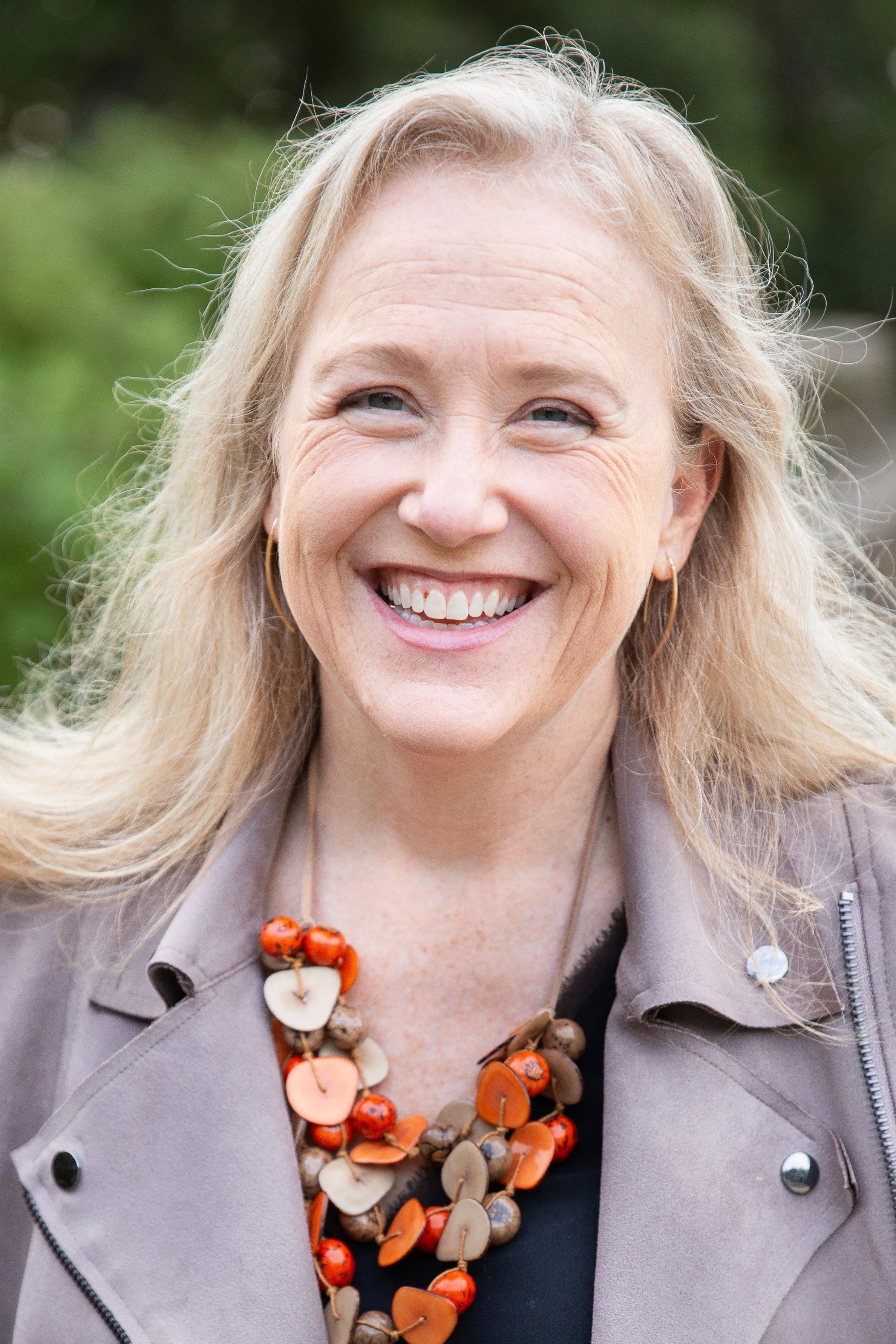 Betsy smiling at the camera wearing a colorful necklace and a neutral colored jacket.