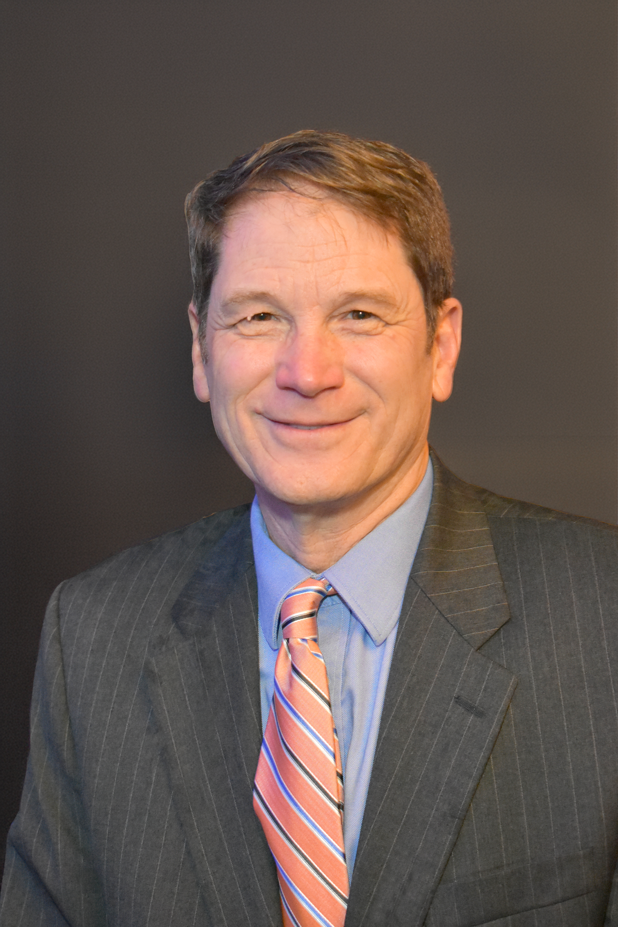 Paul smiling at the camera wearing a dark suit with a light blue shirt and striped tie.