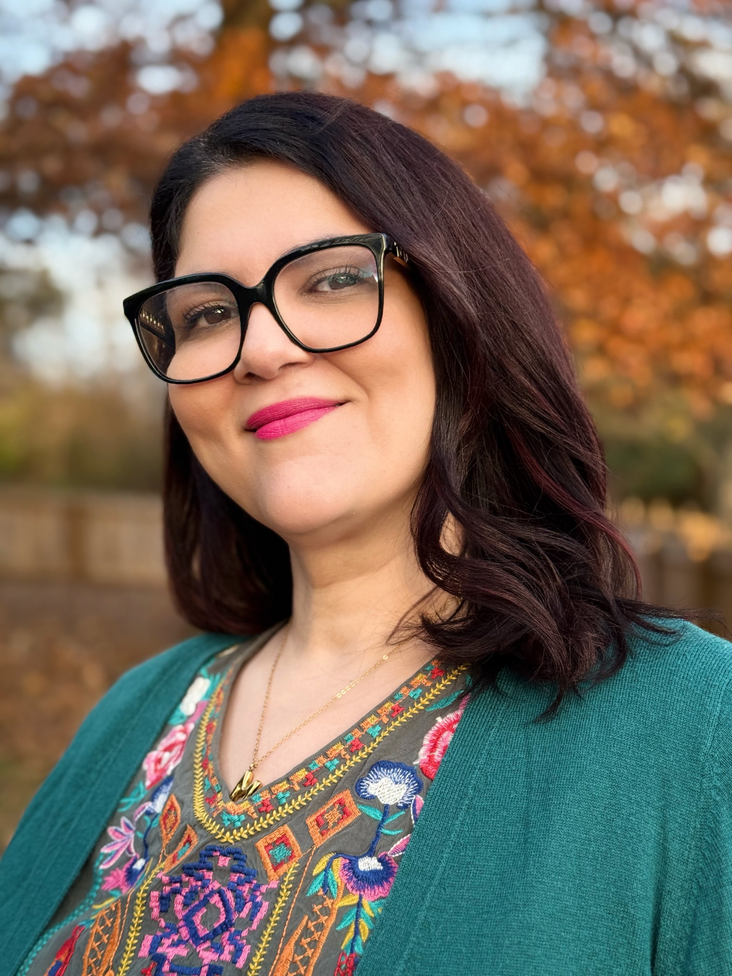Mariel smiling at the camera wearing black glasses and a colorful top with a teal cardigan.
