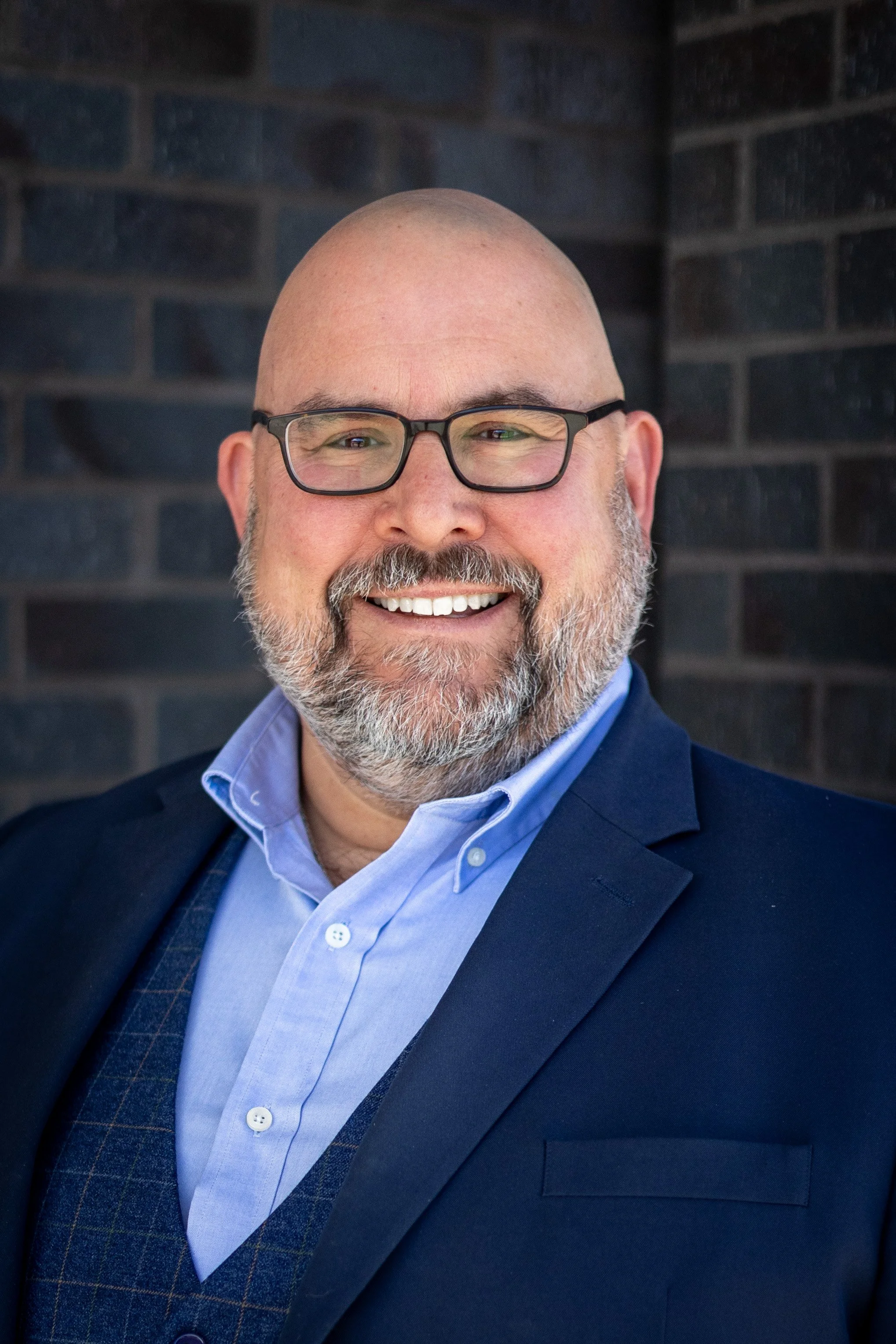 Tom smiling at the camera wearing glasses and a blue suit.