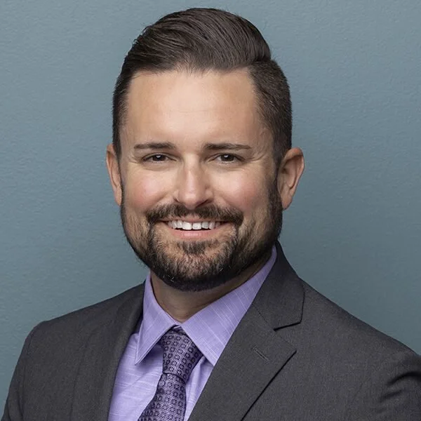 Josh smiling at the camera wearing a dark suit with a purple shirt and tie.