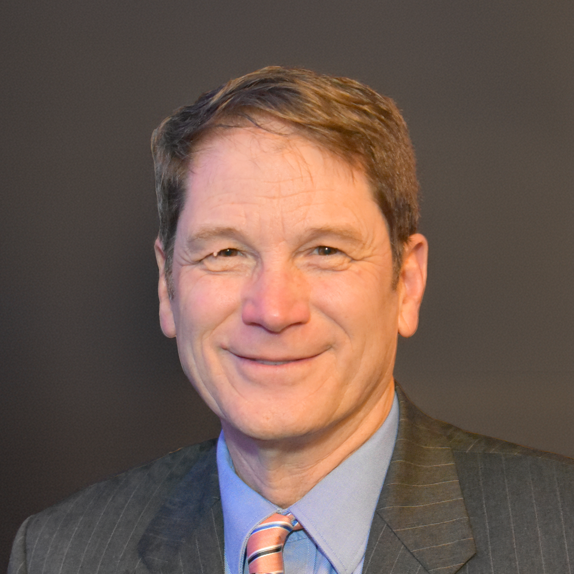 Paul smiling at the camera wear a dark suit with a light blue dress shirt and a pink striped tie.
