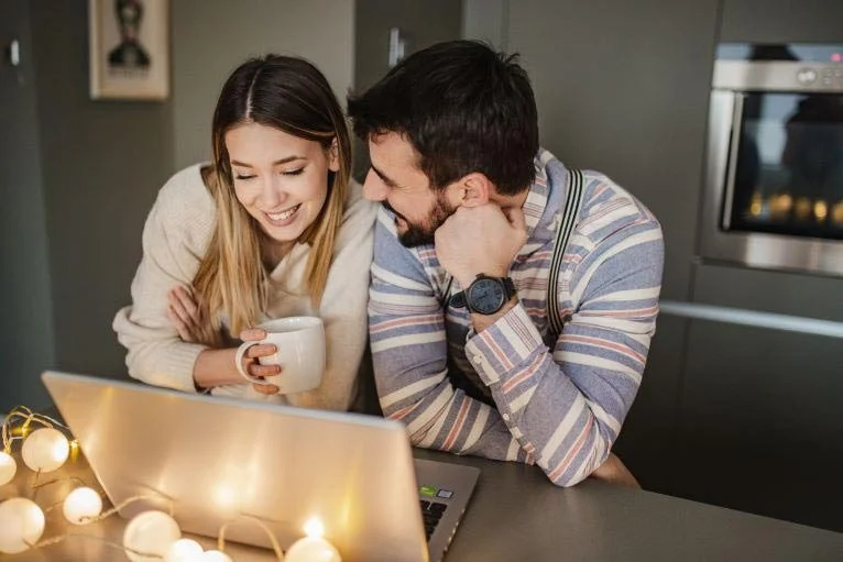 Photograph of a cozy couple in a kitchen. The woman is holding a mug and looking at a laptop, while the man is resting his head on his hand and staring at the woman. They're smiling at each other.