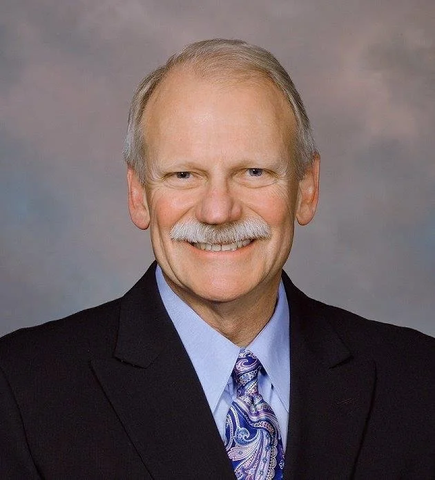 Kim smiling at the camera wearing a dark suit with a light dress shirt and tie.