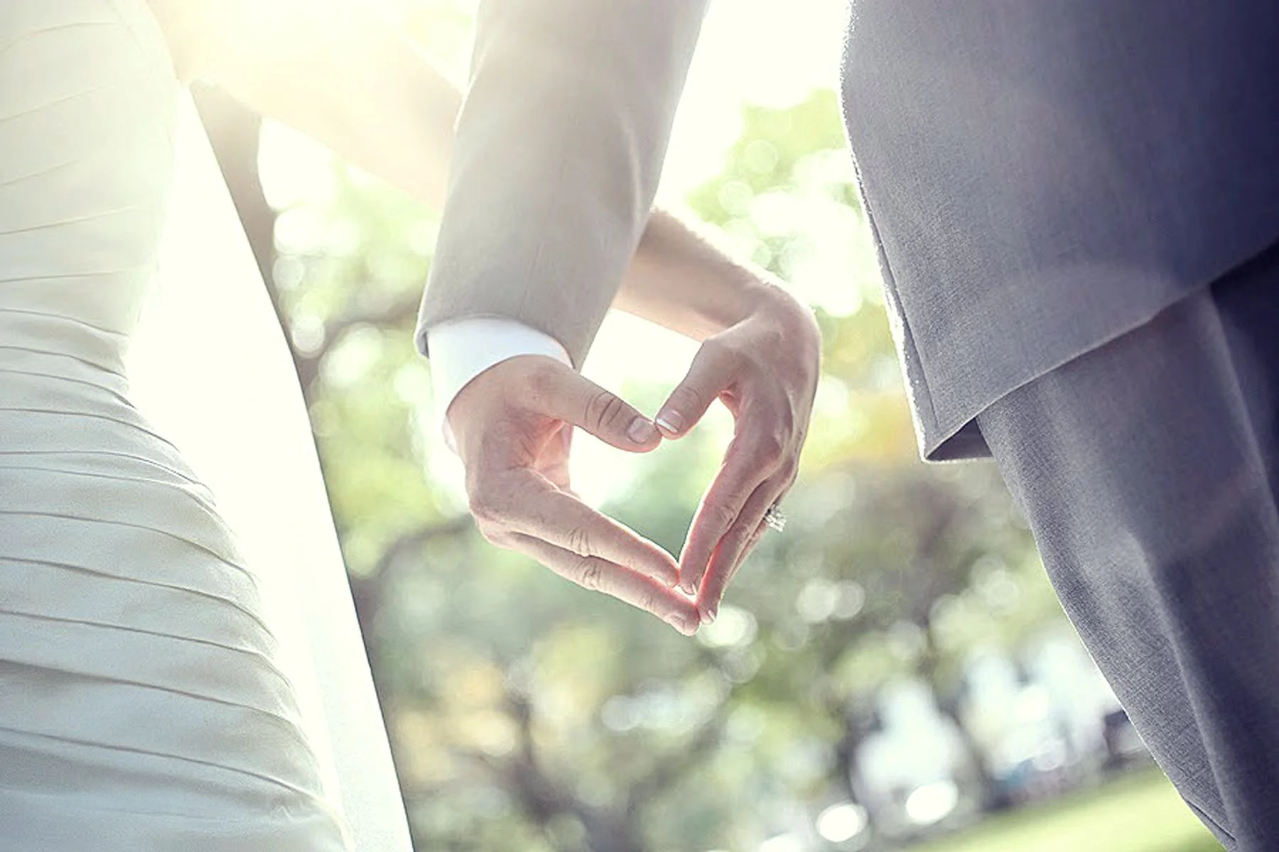 A couple dressed in formal attire making a heart shape with their hands outdoors.