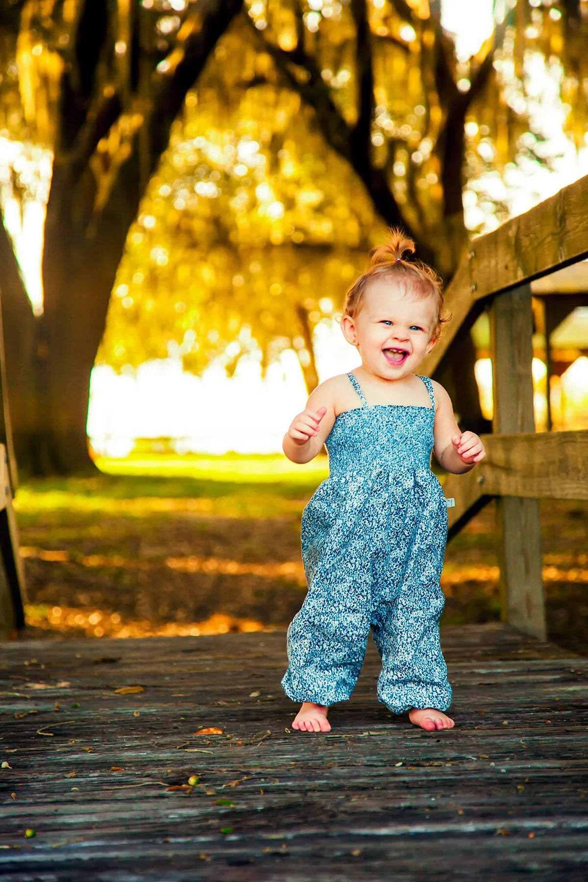 A young girl with a top knot hairstyle, wearing a blue floral jumpsuit, smiling and walking barefoot on a wooden bridge surrounded by trees at sunset.