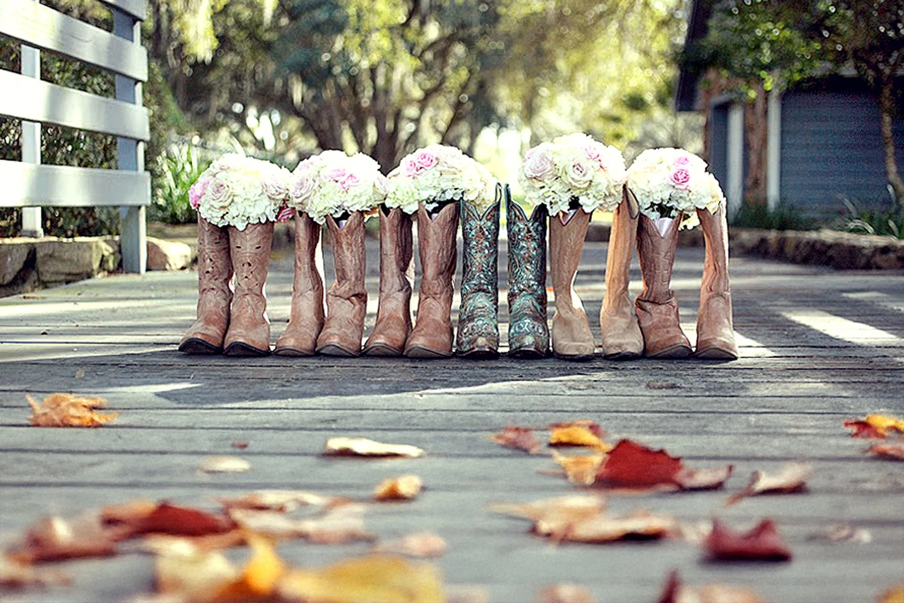 A row of cowboy boots filled with bouquets of pink and white flowers, lined up on a wooden walkway outdoors with trees and a house in the background, and fallen autumn leaves on the ground.