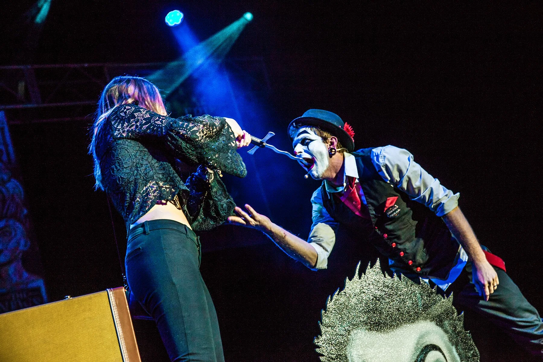 A performer with clown makeup and costume confronts a woman on stage, holding a microphone near her face with theatrical lighting overhead.