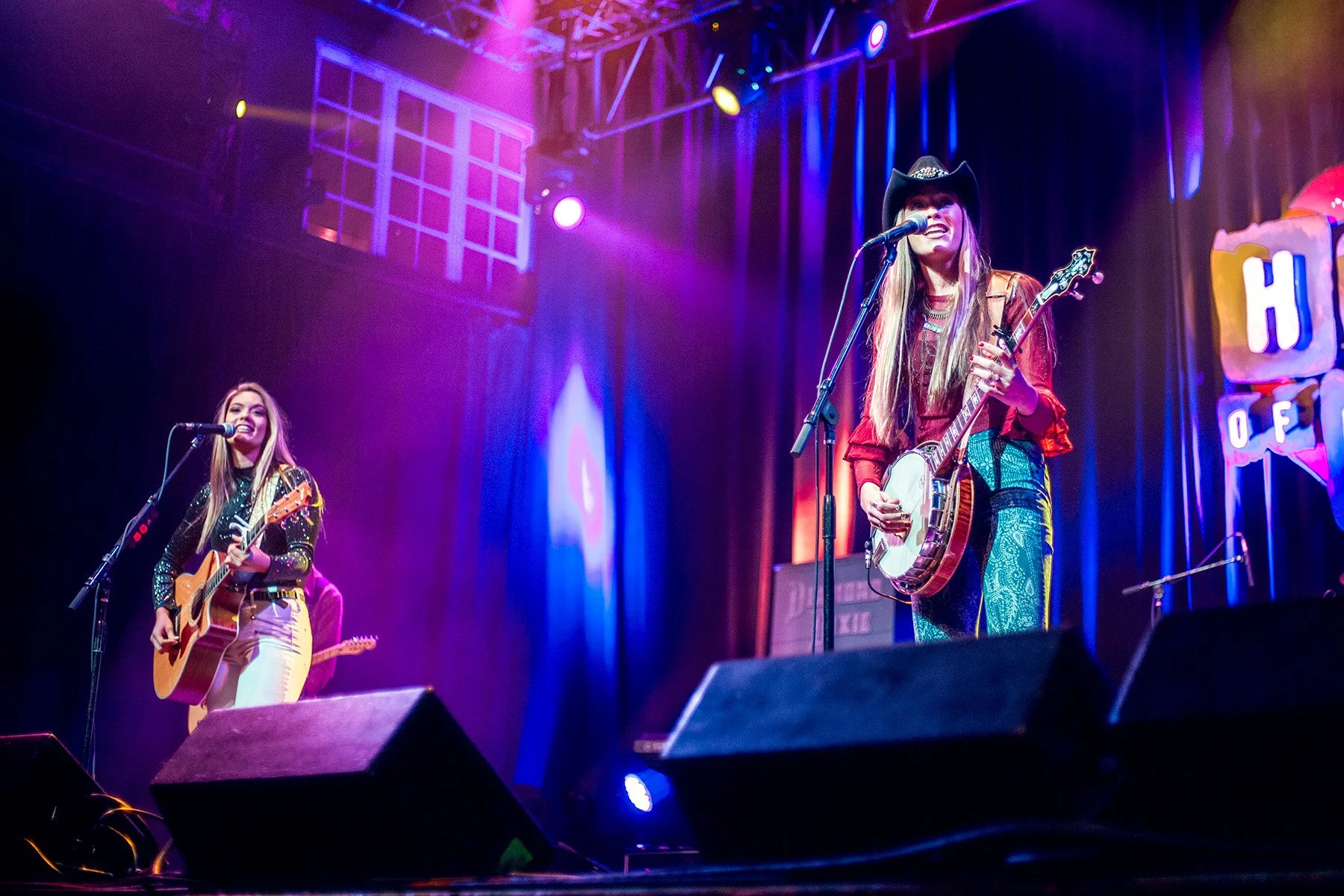 Two women performing on stage with guitars, colorful stage lighting, and a neon sign in the background reading House of Blues.