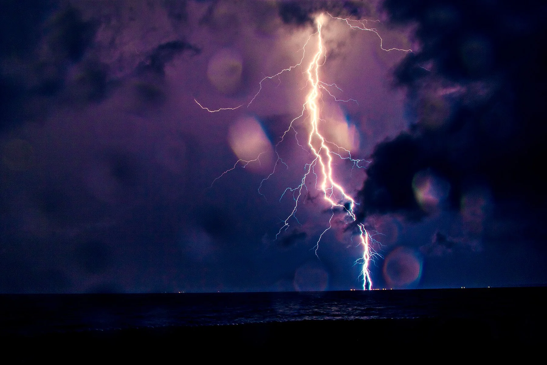 Bright lightning bolt striking the ocean during a thunderstorm at night with dark clouds in the sky.