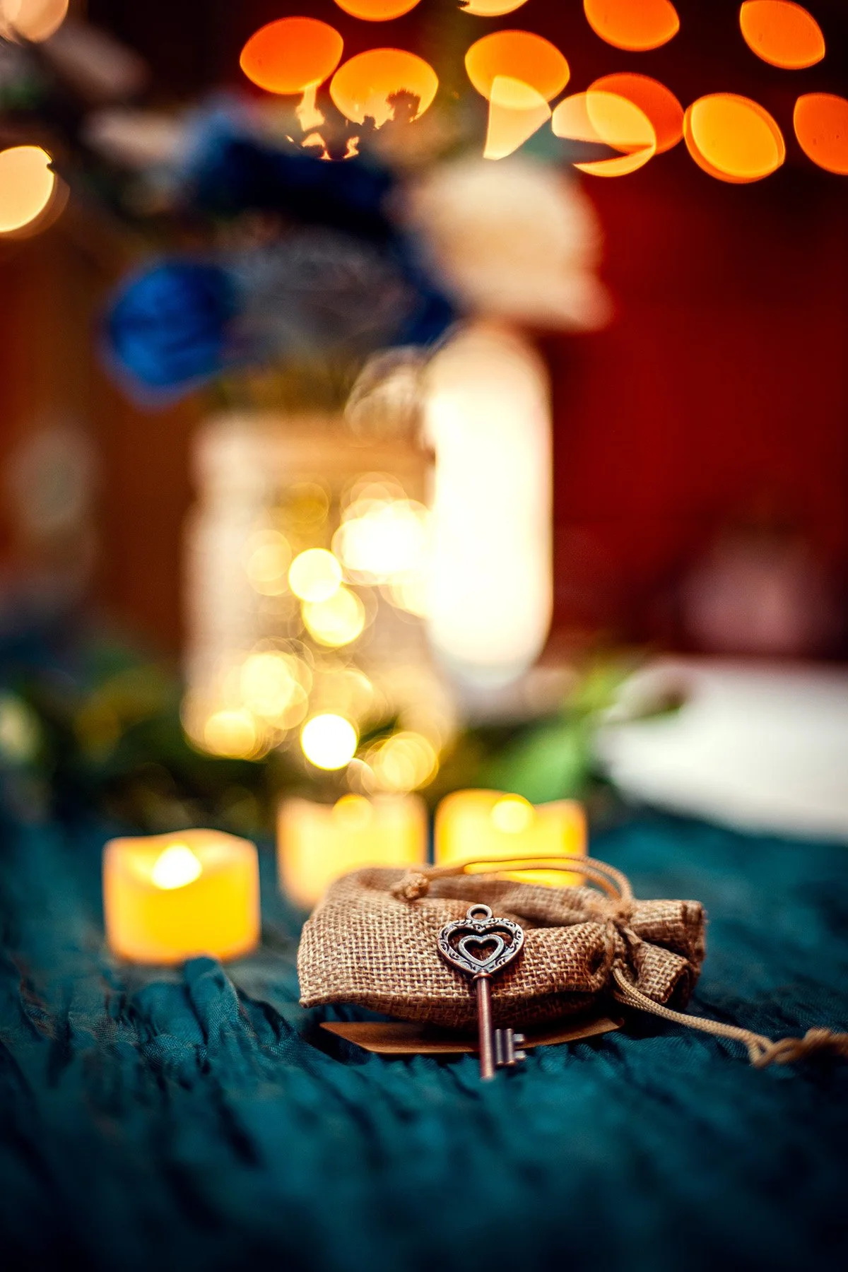 A small, ornate key with a heart-shaped top is placed on a textured brown fabric pouch, surrounded by candle lights and blurred colorful lights in the background.