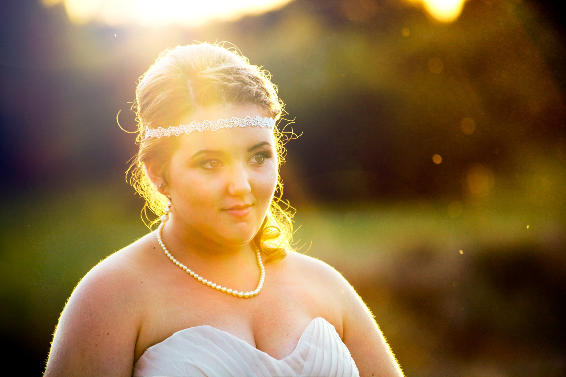 A woman in a white strapless dress with a pearl necklace and headband, outdoors during sunset, with sunlight behind her.