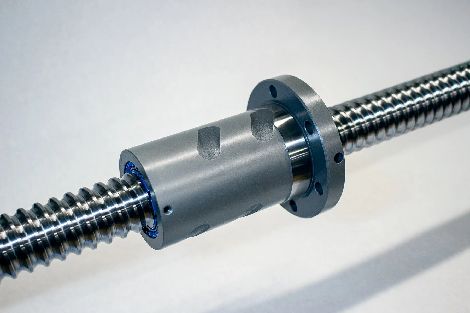 Close-up of a precision mechanical screw spindle with a metallic body, mounted in a machine component, with a light grey background.