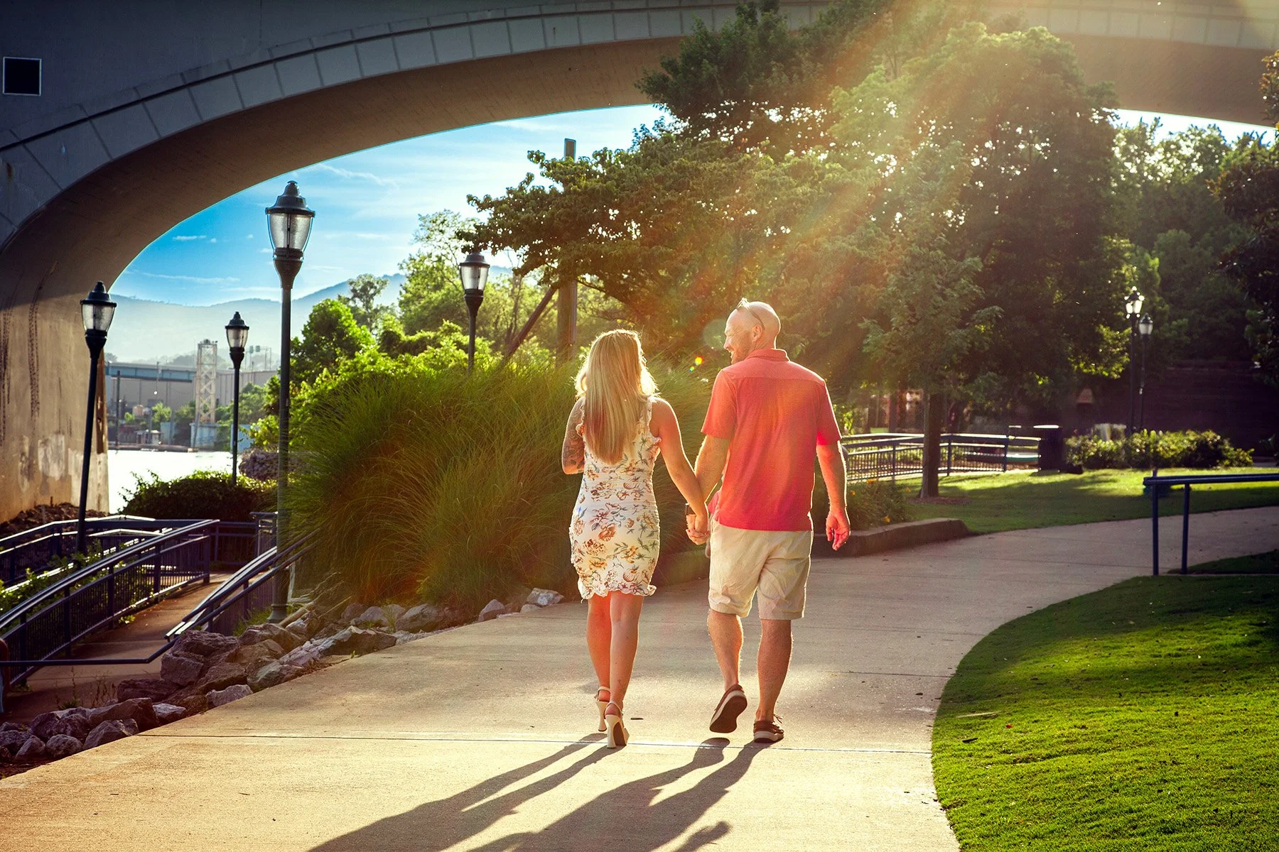 A couple walking hand in hand on a paved path in a park during sunset, with trees, grass, and street lamps around them.