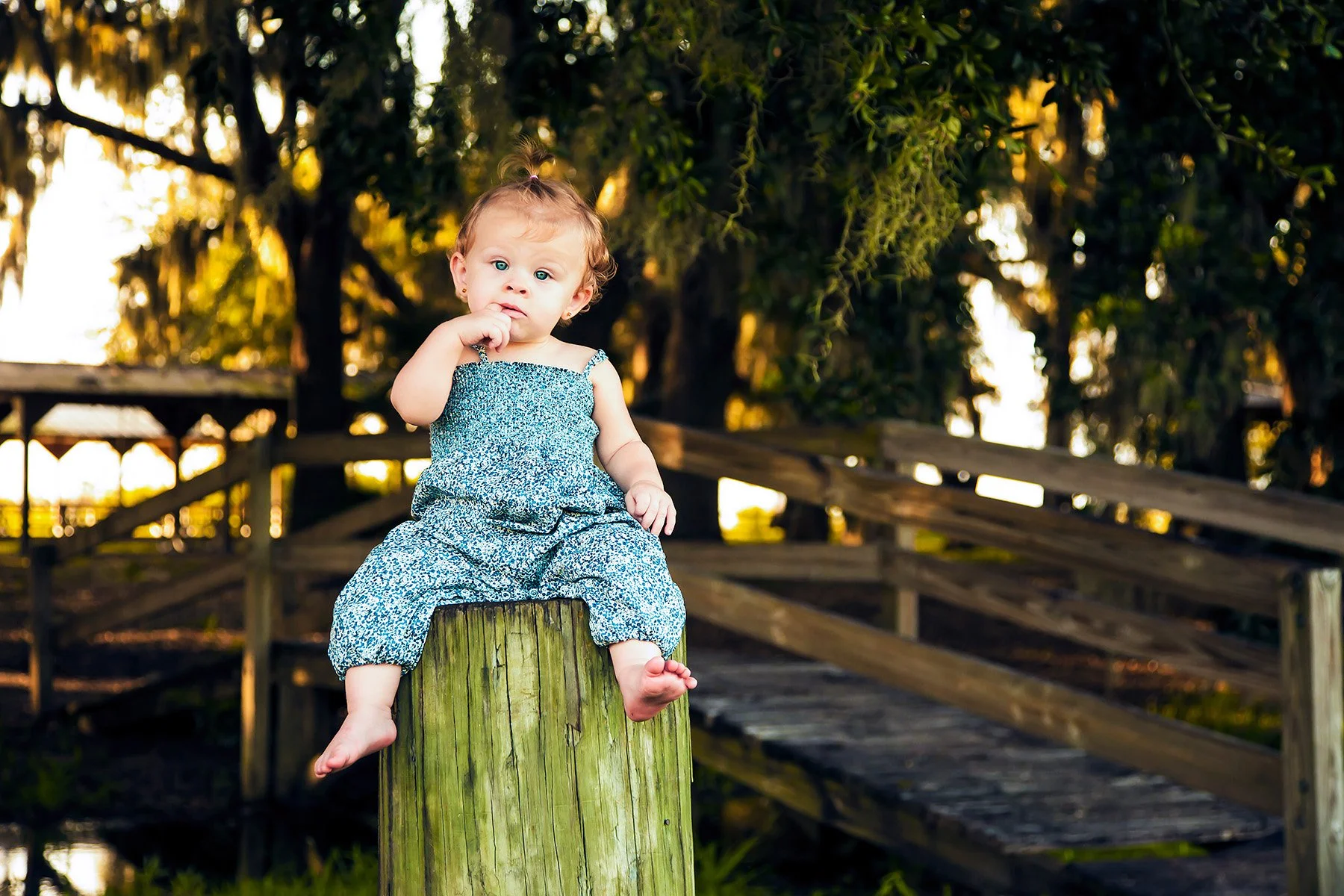 Young girl with blonde hair tied up in a small ponytail, wearing a blue patterned sleeveless jumpsuit, sitting on a wooden post outdoors with trees in the background during sunset.