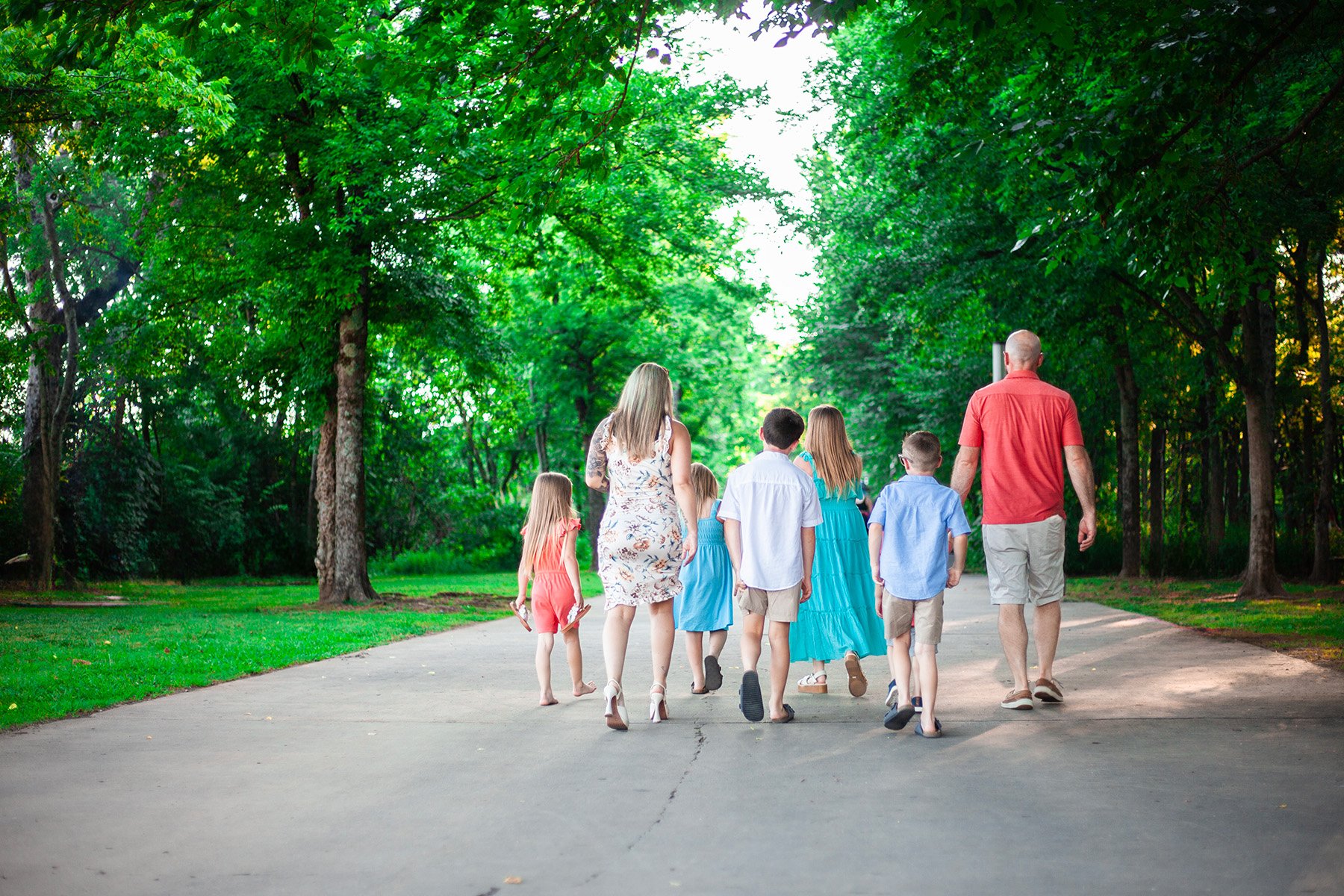 A family walking together on a pathway in a park surrounded by lush green trees, wearing casual summer clothing.