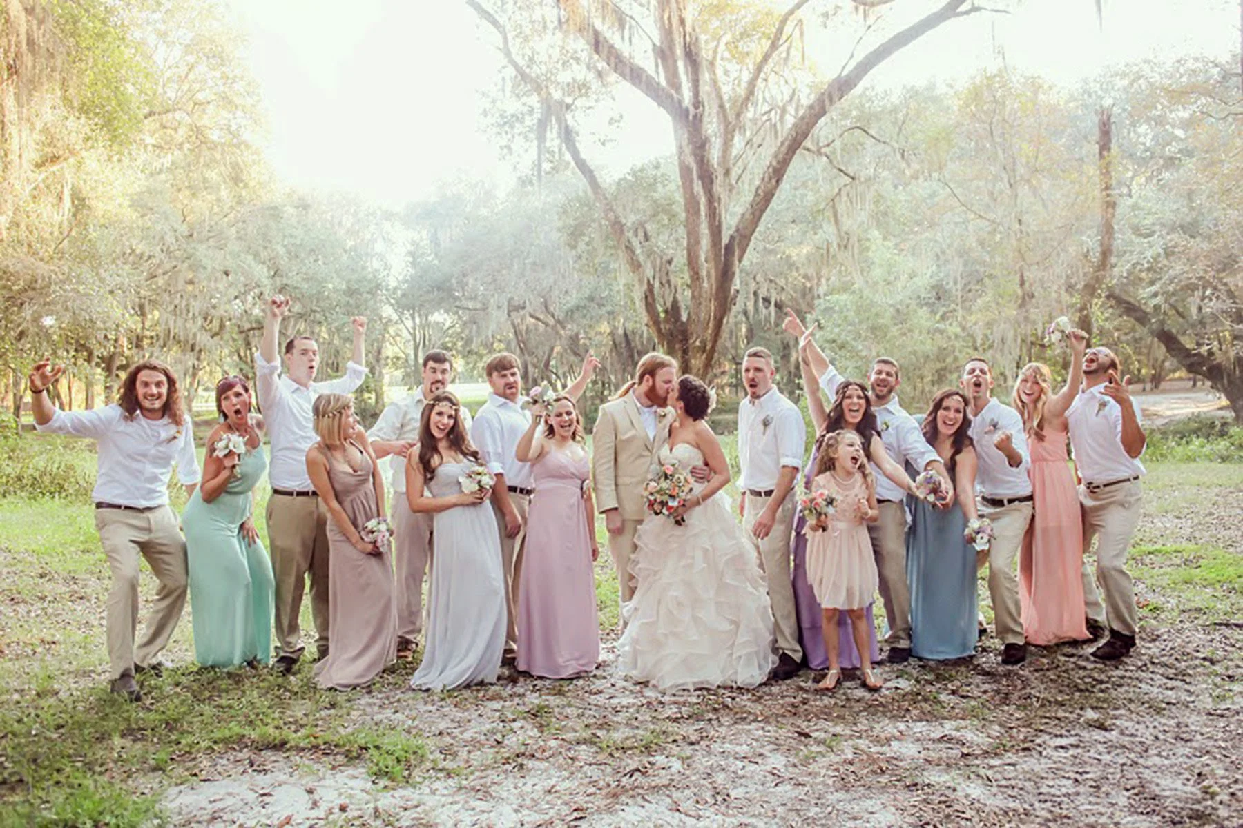A group of people, including a bride and groom, celebrating outdoors in a park with trees, smiling and raising their hands.