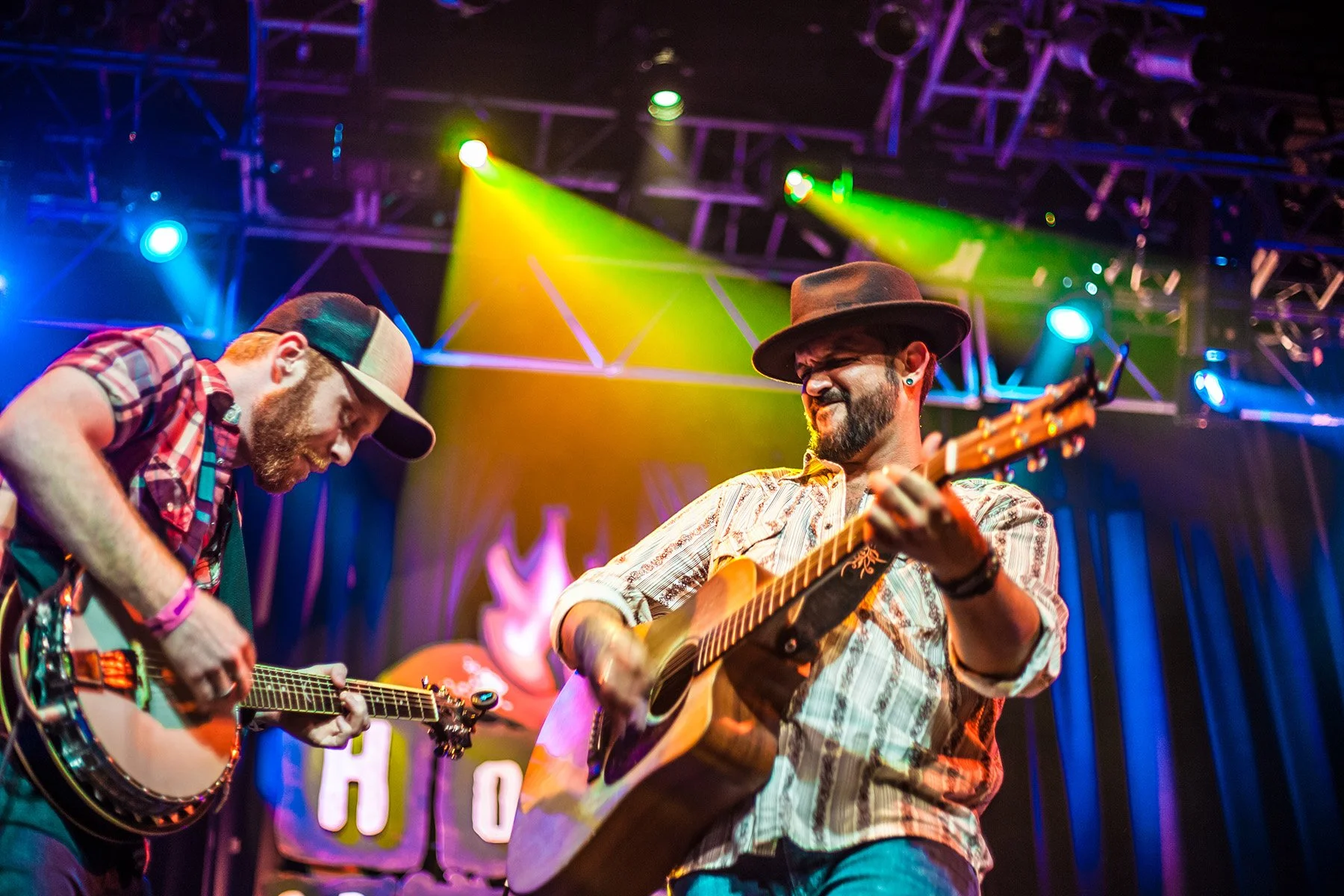 Two musicians playing acoustic guitars on stage with colorful stage lighting, one wearing a plaid shirt and cowboy hat, the other wearing a baseball cap and casual clothing.