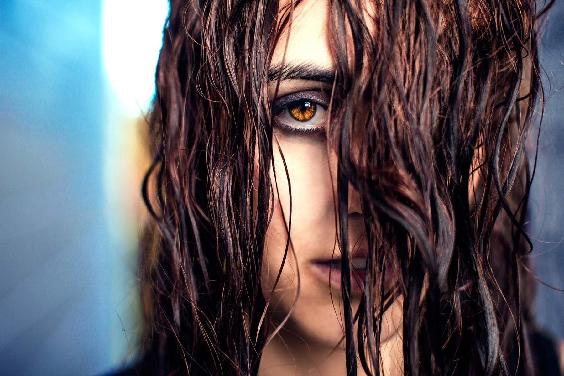 A close-up of a woman's face partially obscured by wet, dark hair, with striking amber eyes looking directly at the camera.
