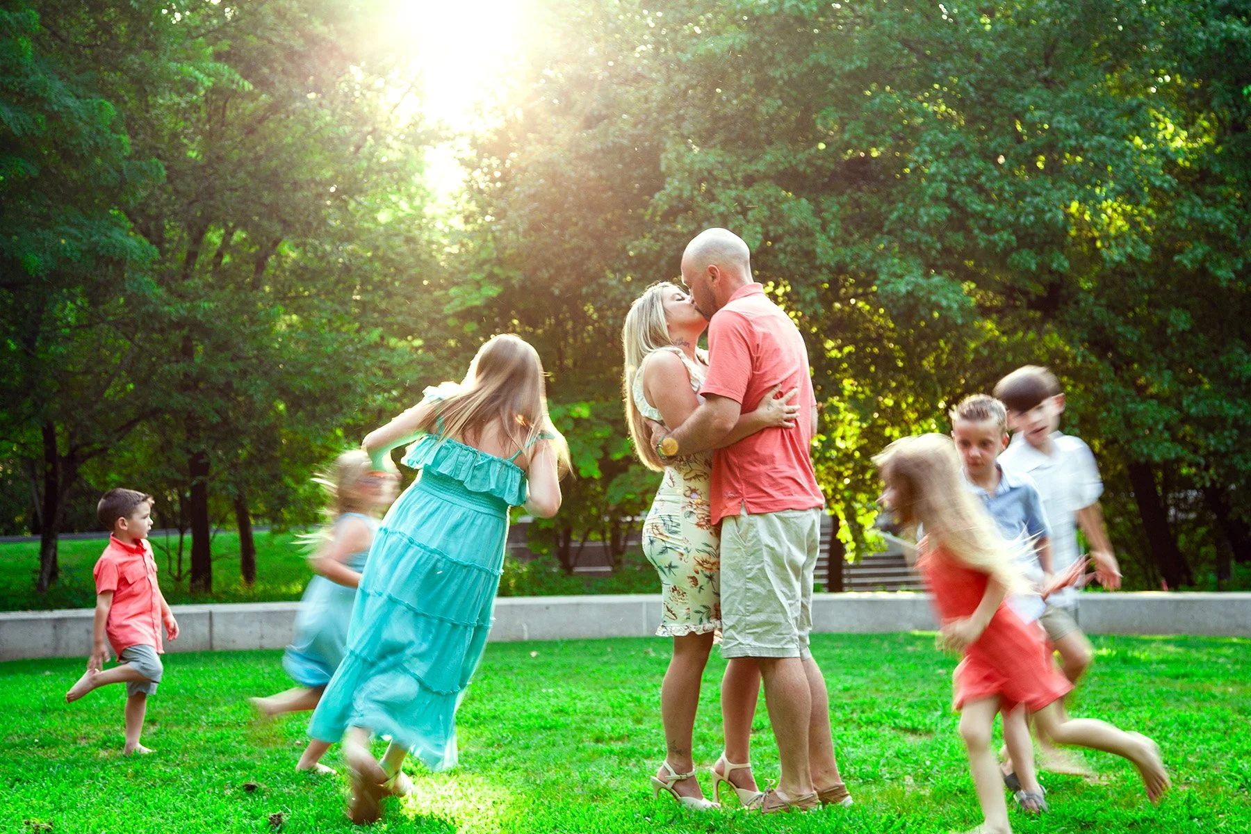 A family kissing in the park with children running around on a sunny day.