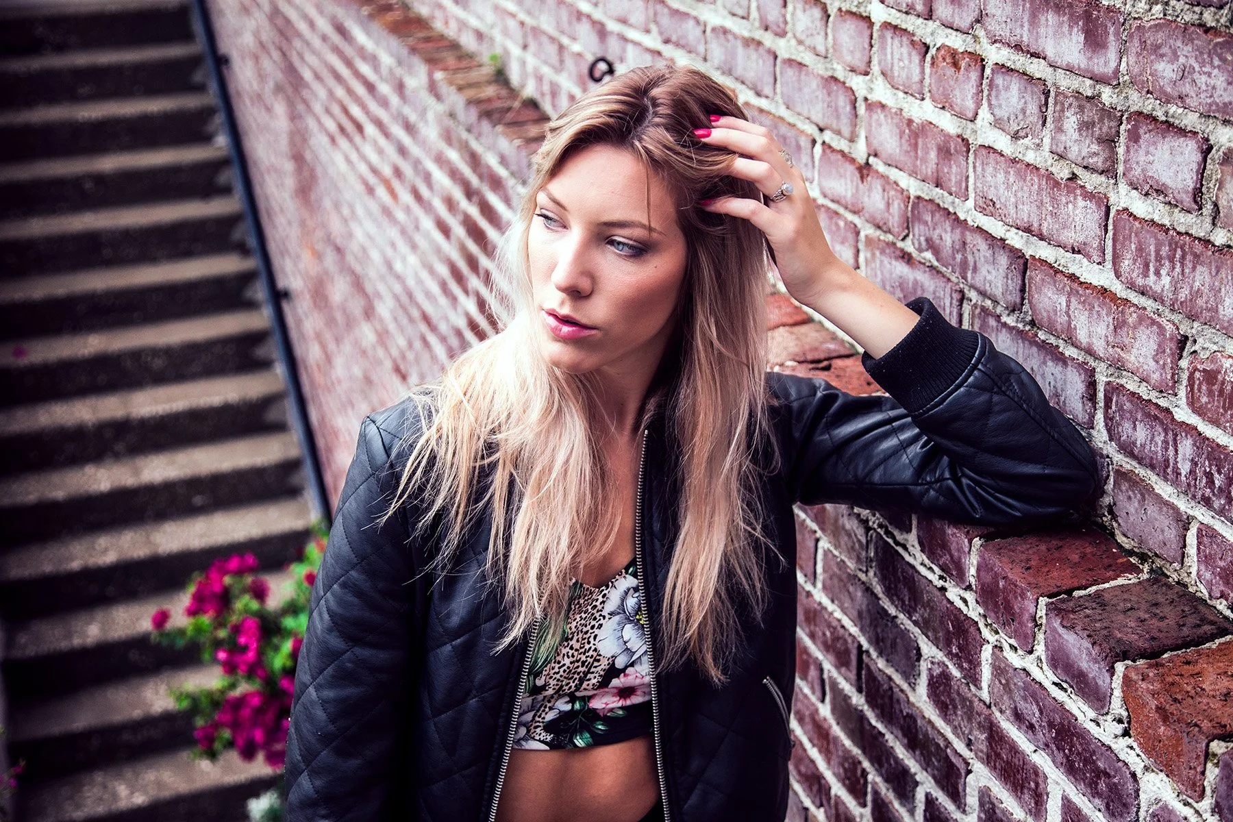 A young woman with long blonde hair, wearing a black jacket and floral top, is sitting against a brick wall while touching her hair, looking thoughtful.