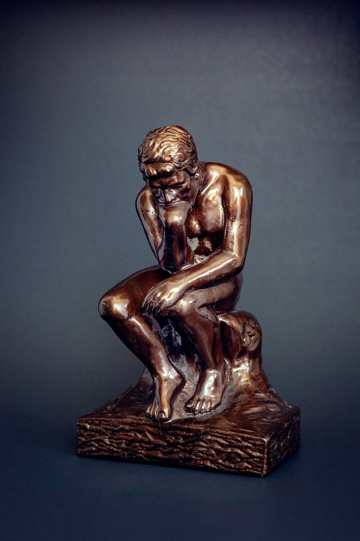 Bronze sculpture of a thinker sitting on a rock with his chin resting on his hand, in a contemplative pose, set against a dark background.