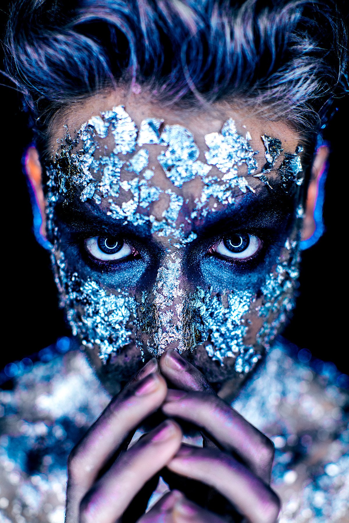 Person with silver foil and makeup covering face, intense blue eyes, and hands pressed together