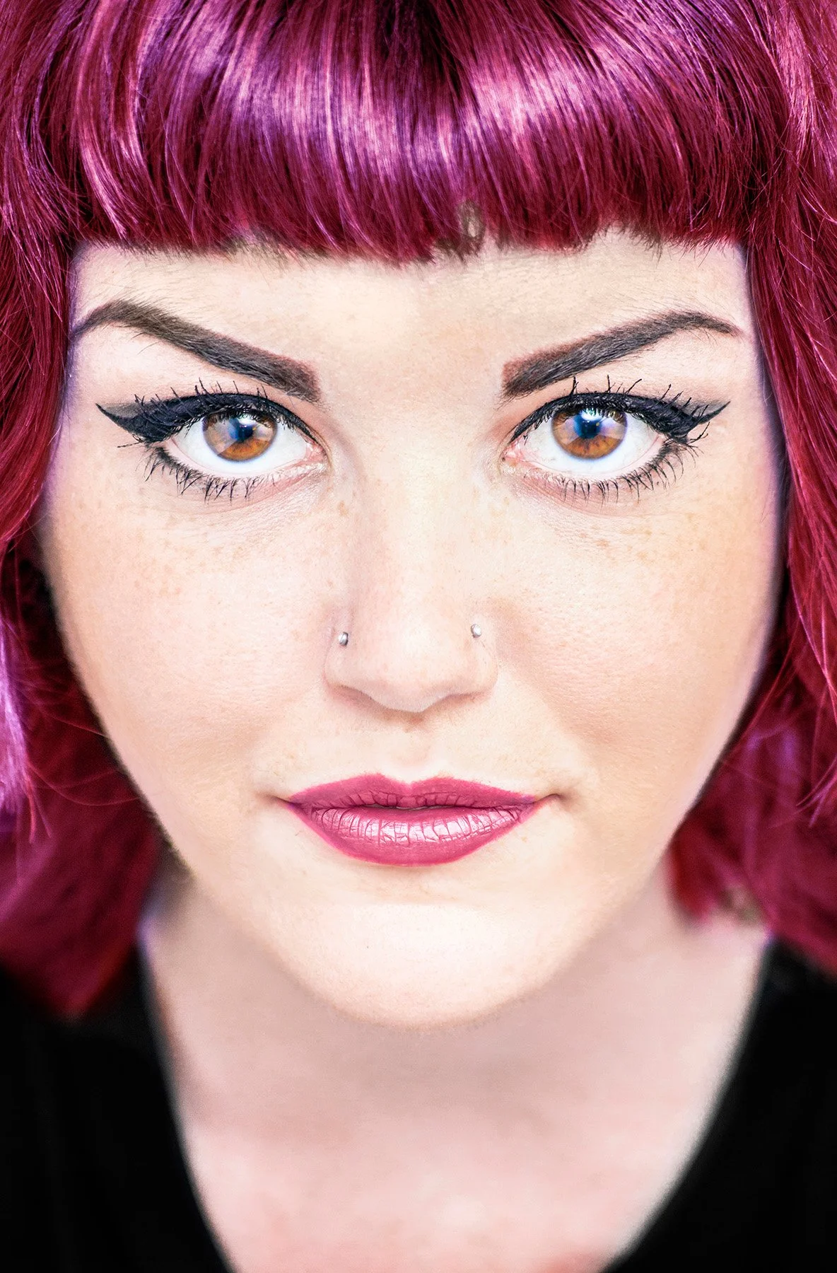 Close-up of a woman with vibrant pink hair, brown eyes with makeup, and a nose piercing, looking directly at the camera.