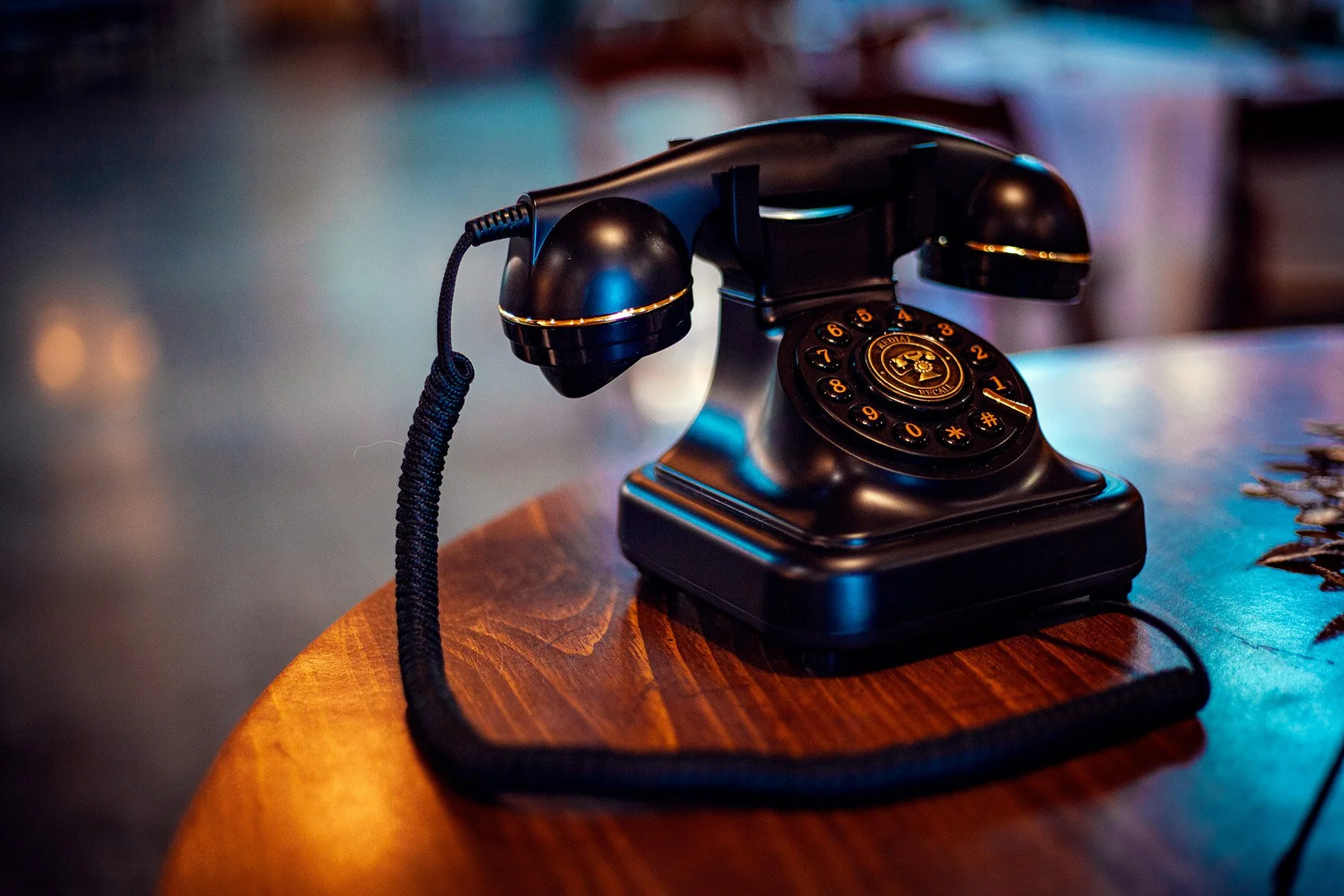 A black, vintage rotary-style telephone with a cord, placed on a wooden table with a natural finish.