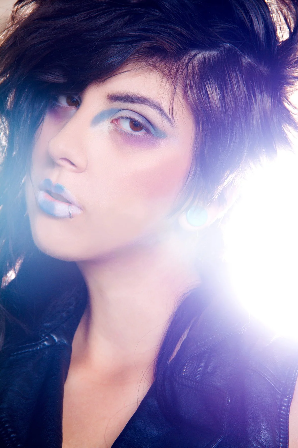Close-up of a woman with dark, styled hair, wearing purple eyeshadow and a black leather jacket, with a bright light source behind her.
