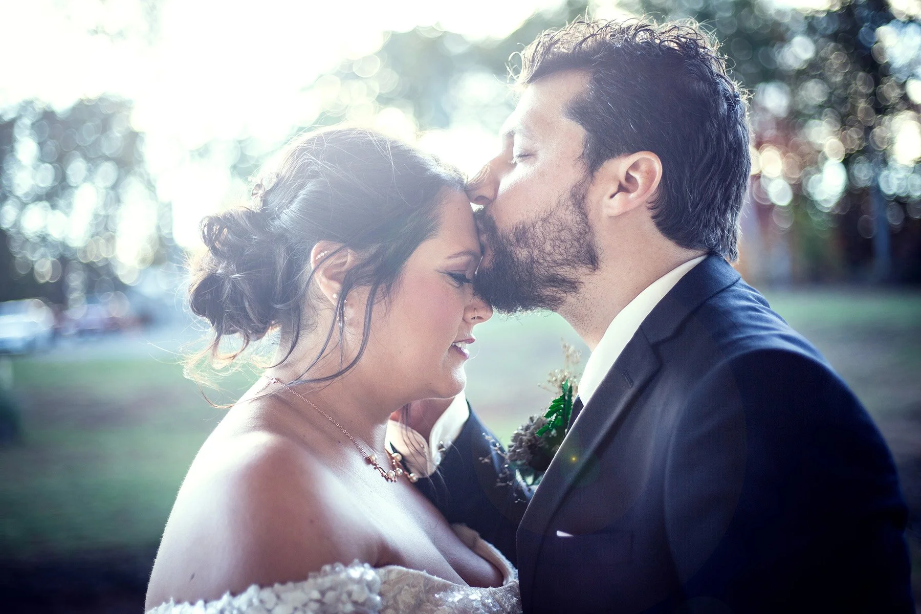 A bride and groom sharing a romantic moment outdoors, with the groom gently kissing the bride's forehead. The bride has her eyes closed and wears a lace wedding dress and necklace, while the groom is in a dark suit with a boutonniere.