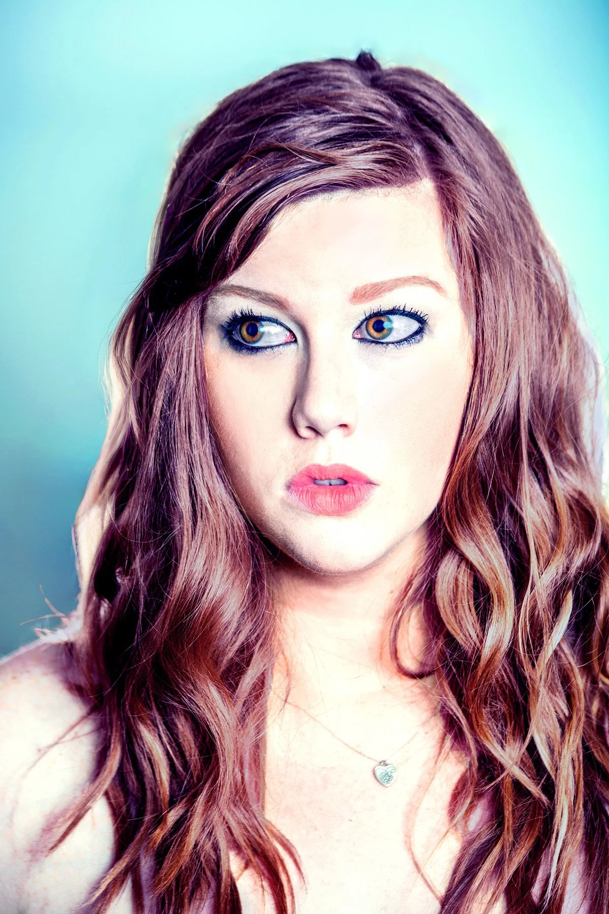 Close-up portrait of a young woman with long wavy reddish-brown hair, blue eyes, light makeup, and a heart-shaped necklace against a soft teal background.