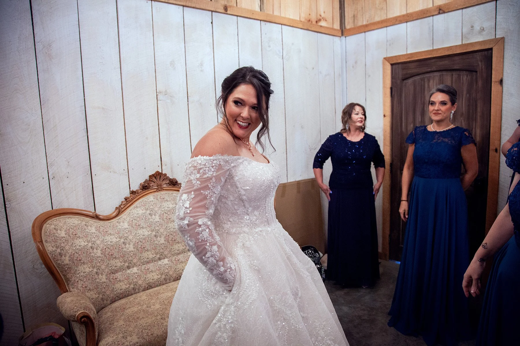 A bride in a white wedding dress smiling, with three women in blue dresses in the background, inside a wooden room with white panel walls and a floral upholstered vintage sofa.