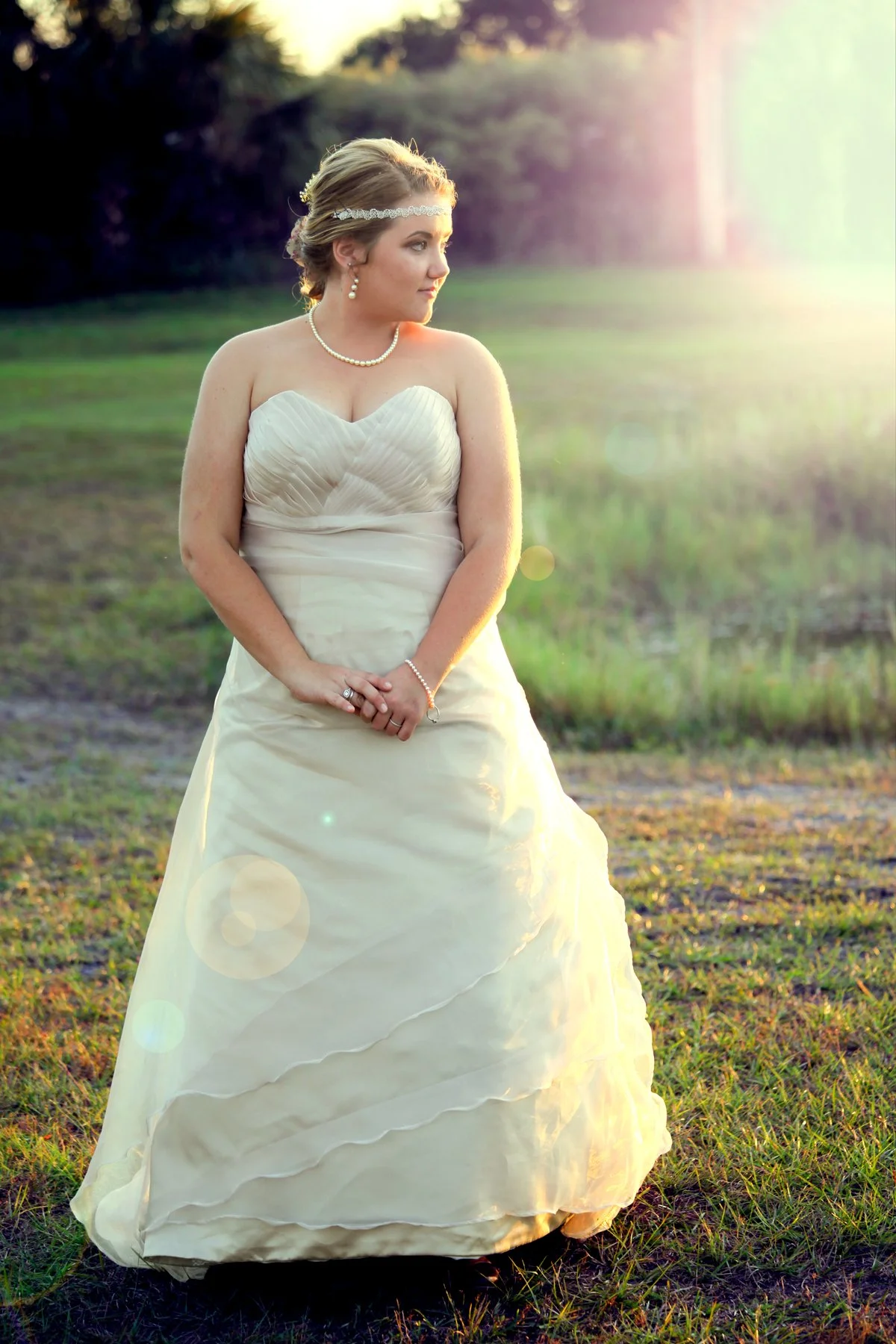 A bride in a strapless cream-colored wedding gown standing outdoors on a grassy field at sunset, with trees in the background and sunlight creating a soft glow.