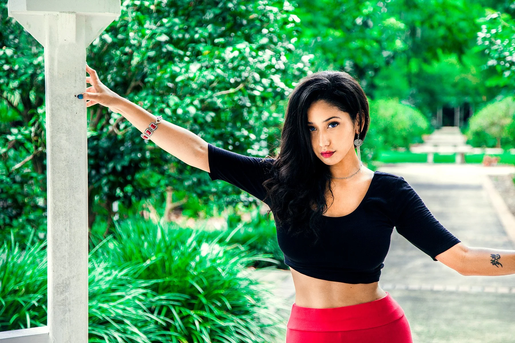 A young woman with long black hair wearing a black crop top and a red skirt, standing outdoors with green trees and foliage in the background, holding onto a pole.