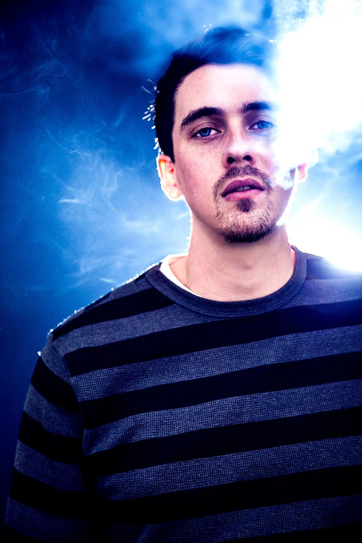 A young man with blue eyes and dark hair, wearing a black and gray striped shirt, stands against a background with bright white and blue lighting, with smoke around him.