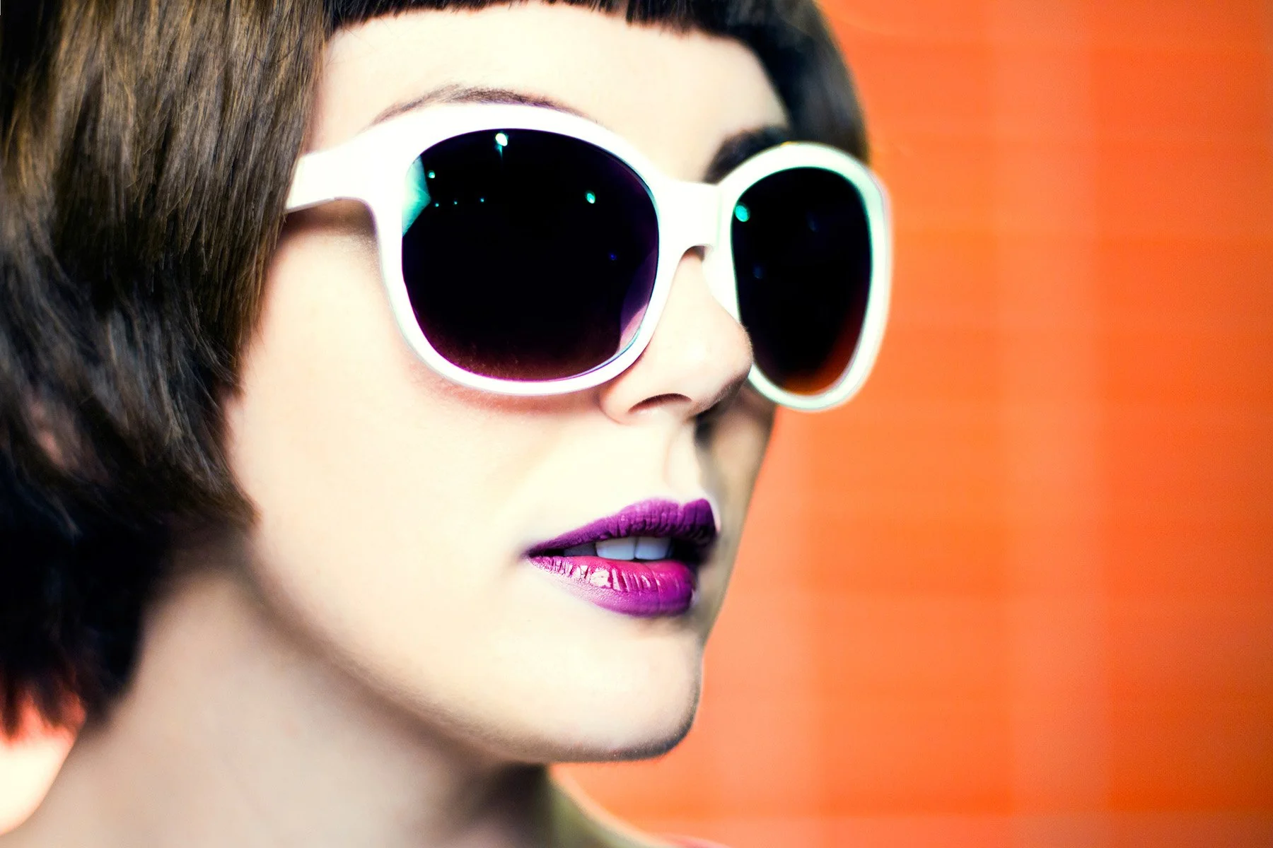 A woman with short dark hair wearing white-framed sunglasses with dark lenses, bright pink lipstick, and lipstick with purple undertones, looking to the side with a blurred orange background.