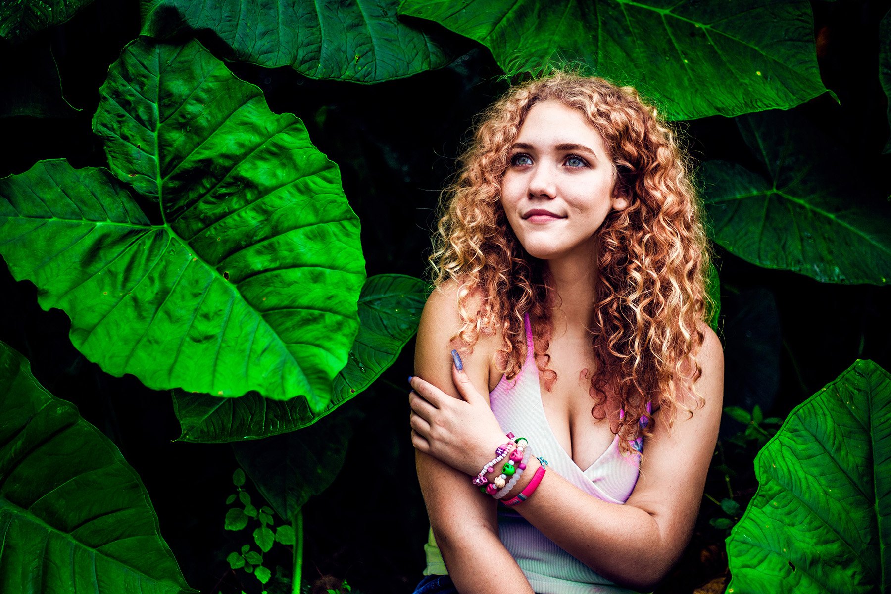 Young woman with curly red hair and blue eyes standing among large green tropical leaves, wearing a light pink tank top and colorful bracelets.