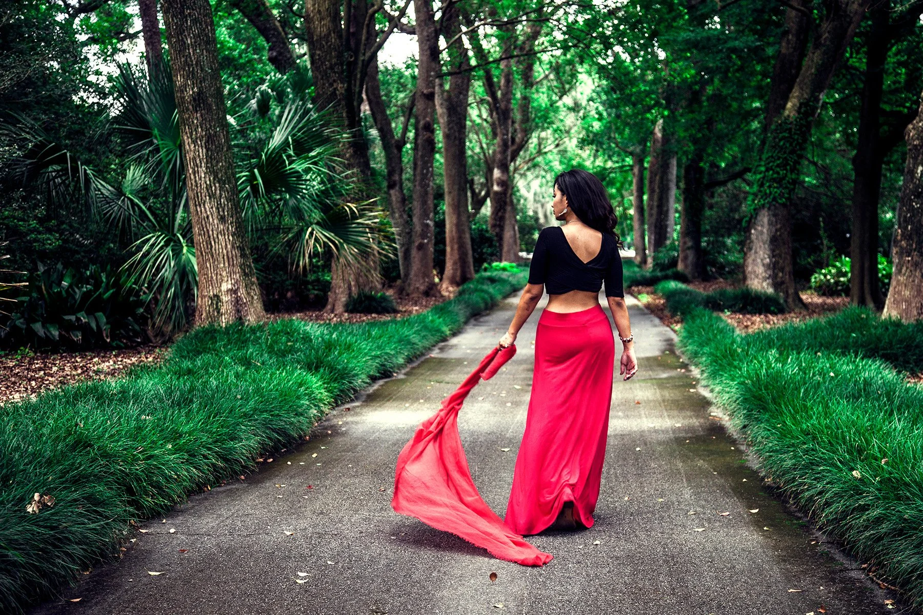 A woman walking down a lush green forest pathway, wearing a black long-sleeved crop top and a flowing red skirt, holding the end of her skirt in her left hand.