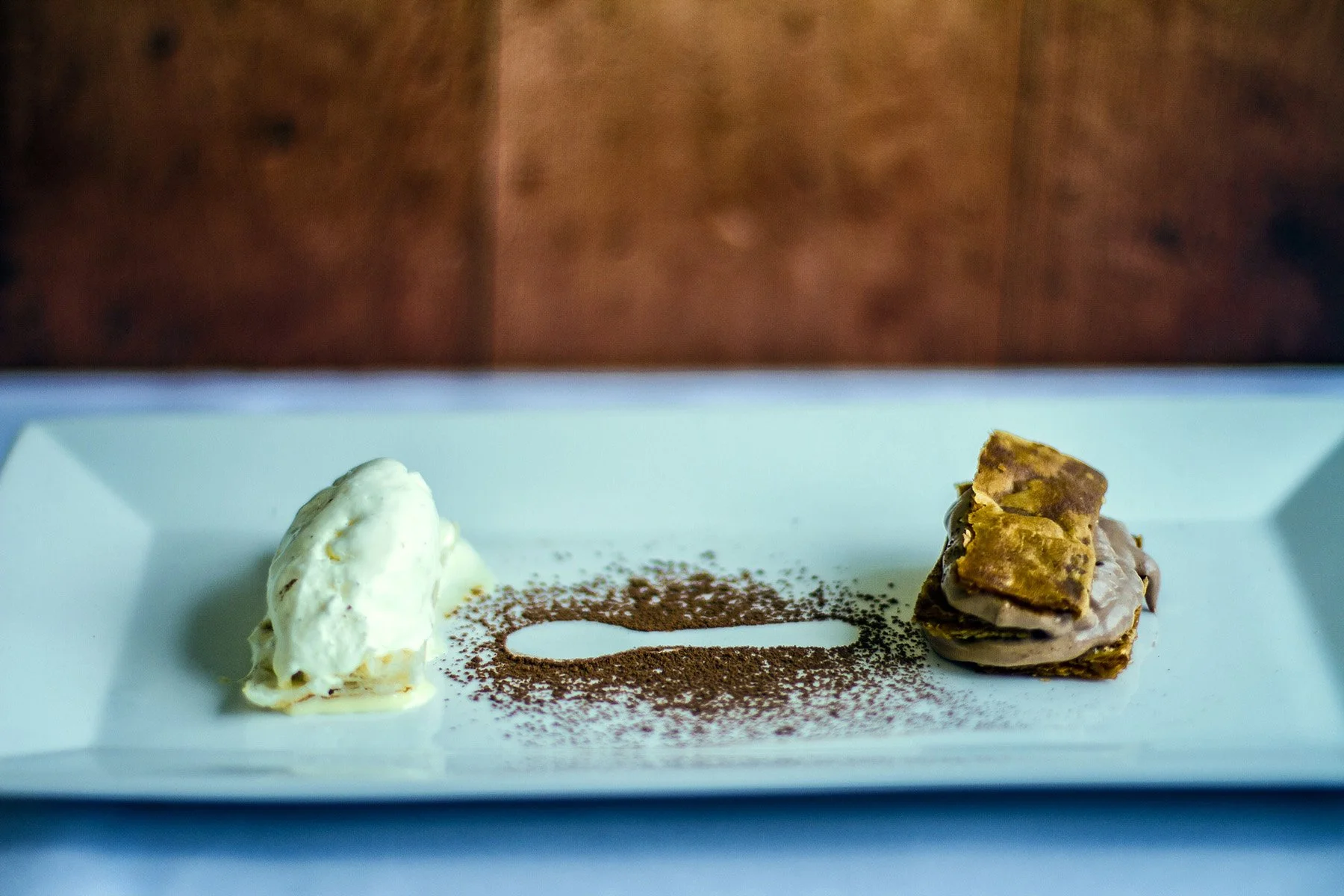 A plated dessert featuring a dollop of whipped cream on the left and a piece of layered chocolate cake on the right, with cocoa powder sprinkled on the white plate.