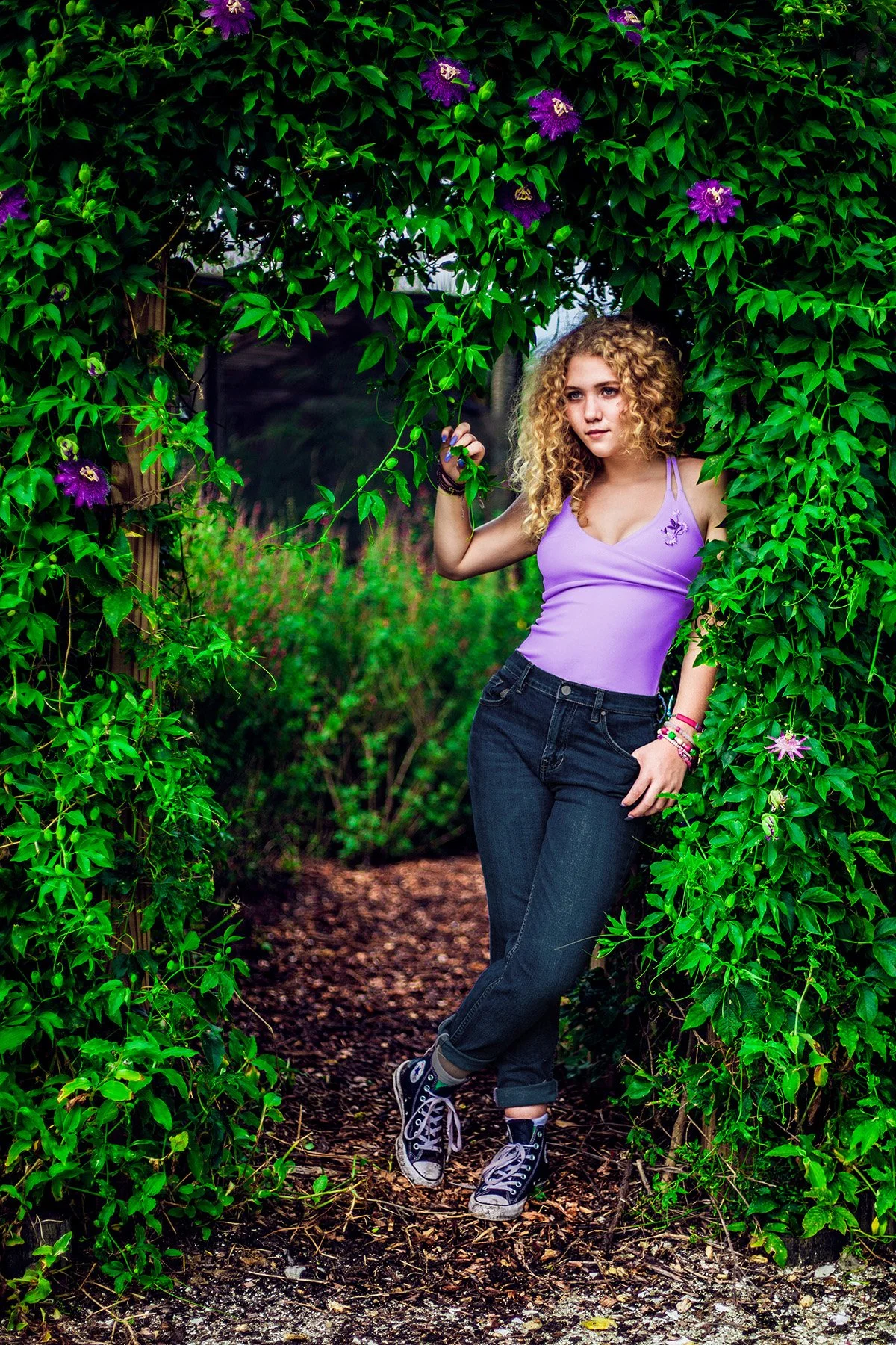 A young woman with curly blonde hair standing in a lush green garden framed by an archway of foliage and purple flowers, wearing a lavender tank top, dark jeans, and black sneakers.