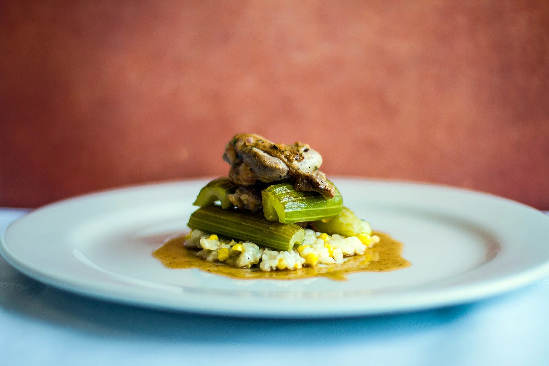 A plated dish featuring a small portion of rice with corn, topped with grilled celery and a piece of grilled chicken, and drizzled with brown gravy.