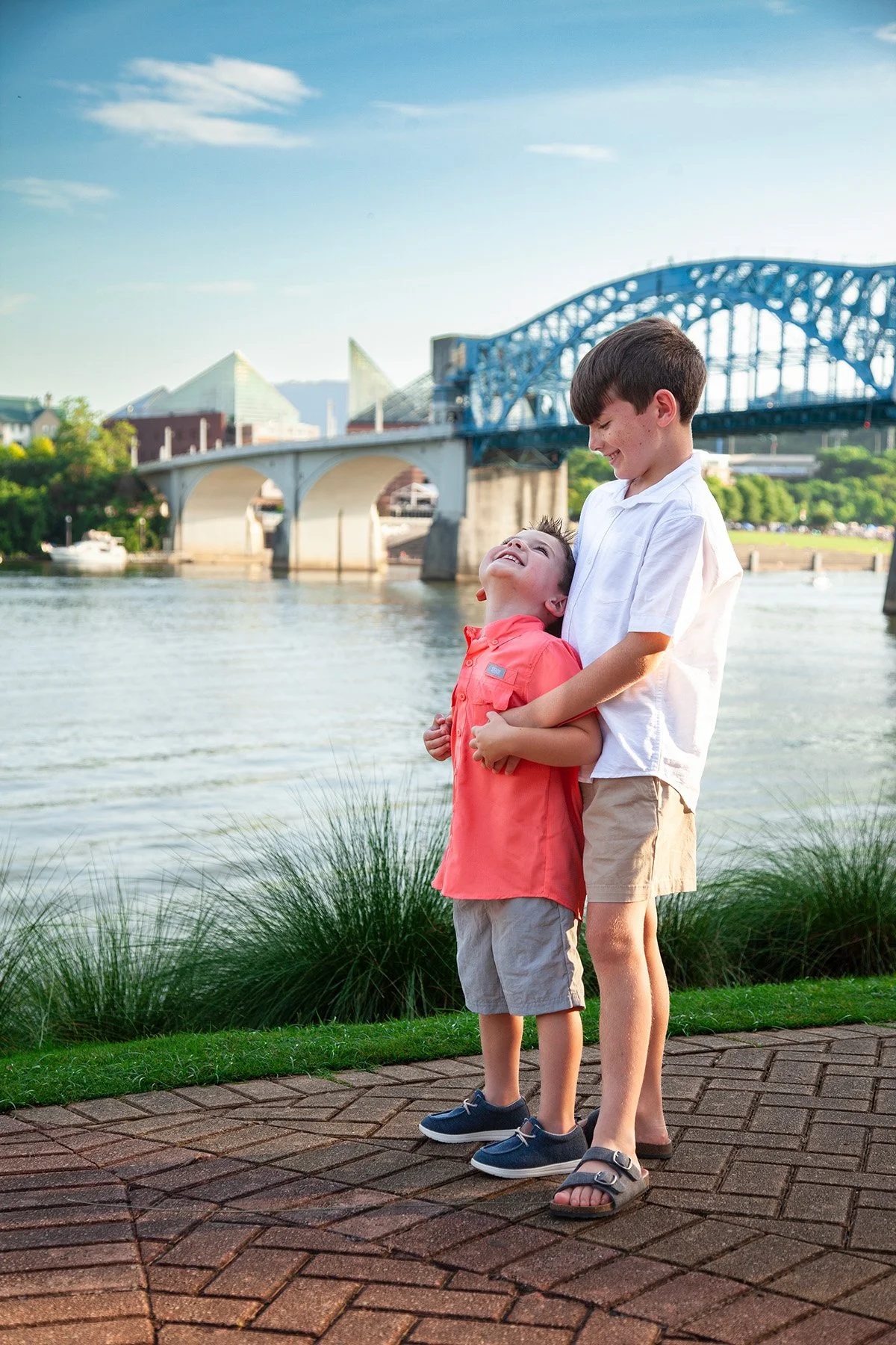 Two boys are standing close by a river, smiling and holding each other. They are outdoors with a bridge and city skyline in the background.
