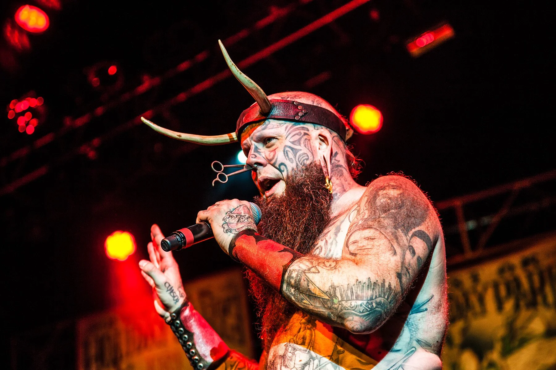 Tattooed man with a beard wearing a Viking helmet with horns, holding a microphone, performing on stage under red lighting.