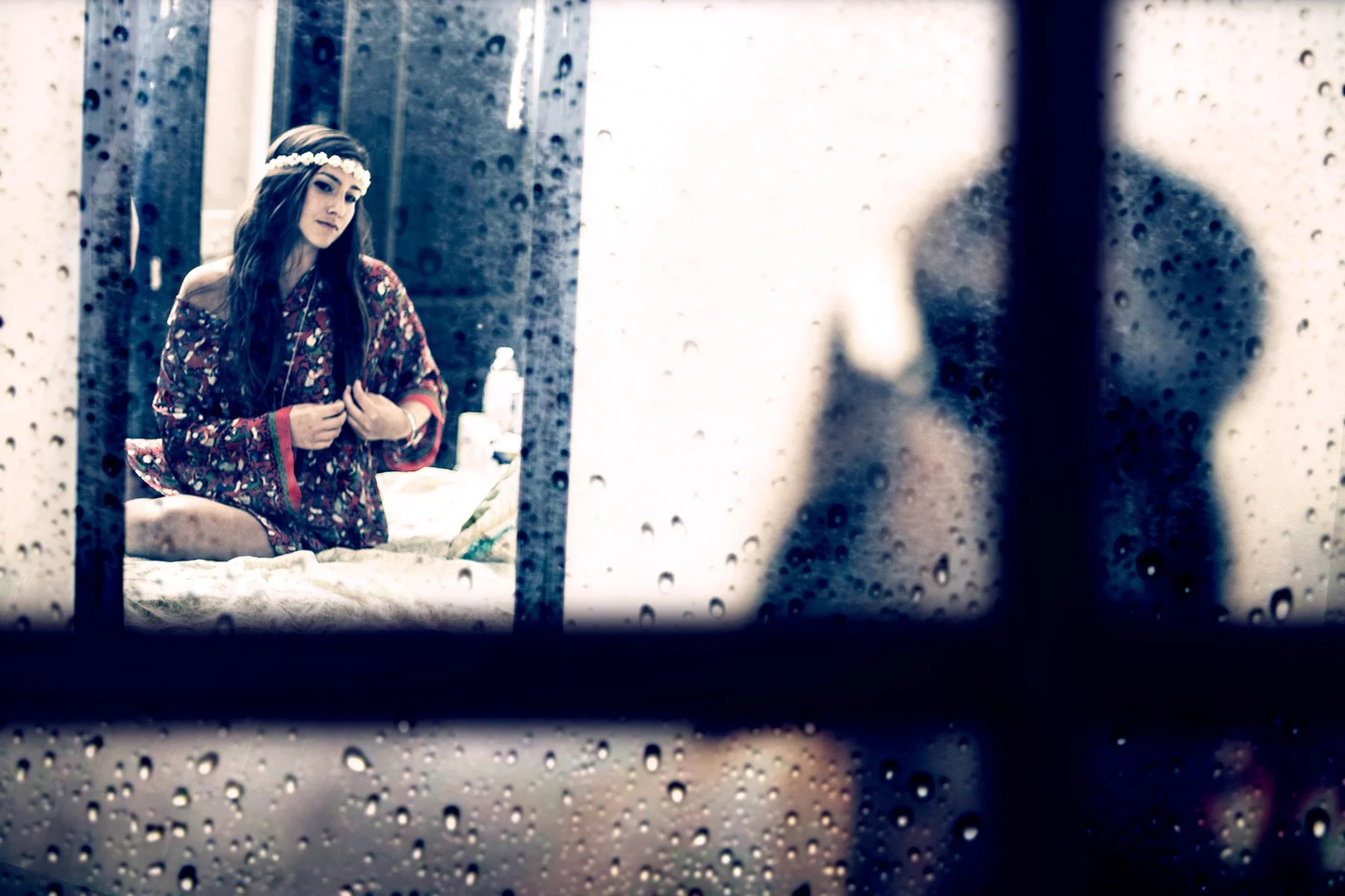 A woman with long dark hair, wearing a floral dress and a flower headband, is seen through a rain-covered window. She appears to be inside a bedroom, sitting on a bed with a slightly pensive expression.